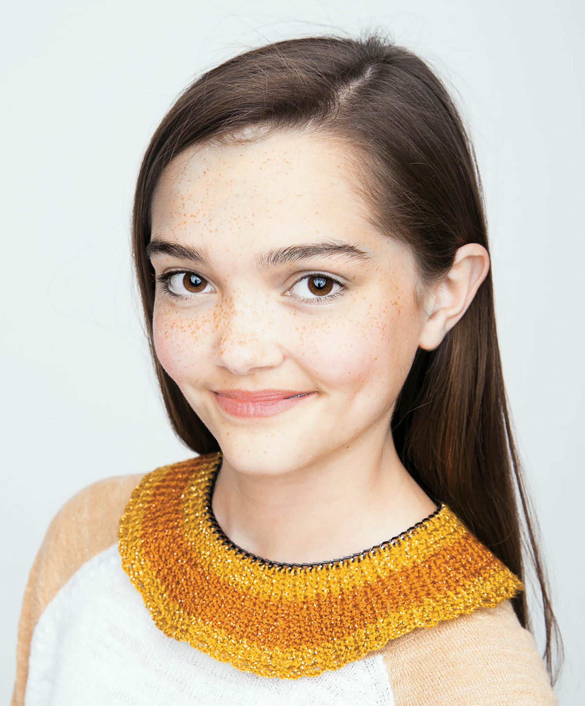 A young woman with straight brown hair and freckles smiles softly, modeling a chunky golden yellow crochet collar from Crochet Collars for Kids by Leisure Arts. She pairs it with a white top and tan sleeves against a plain, light background.