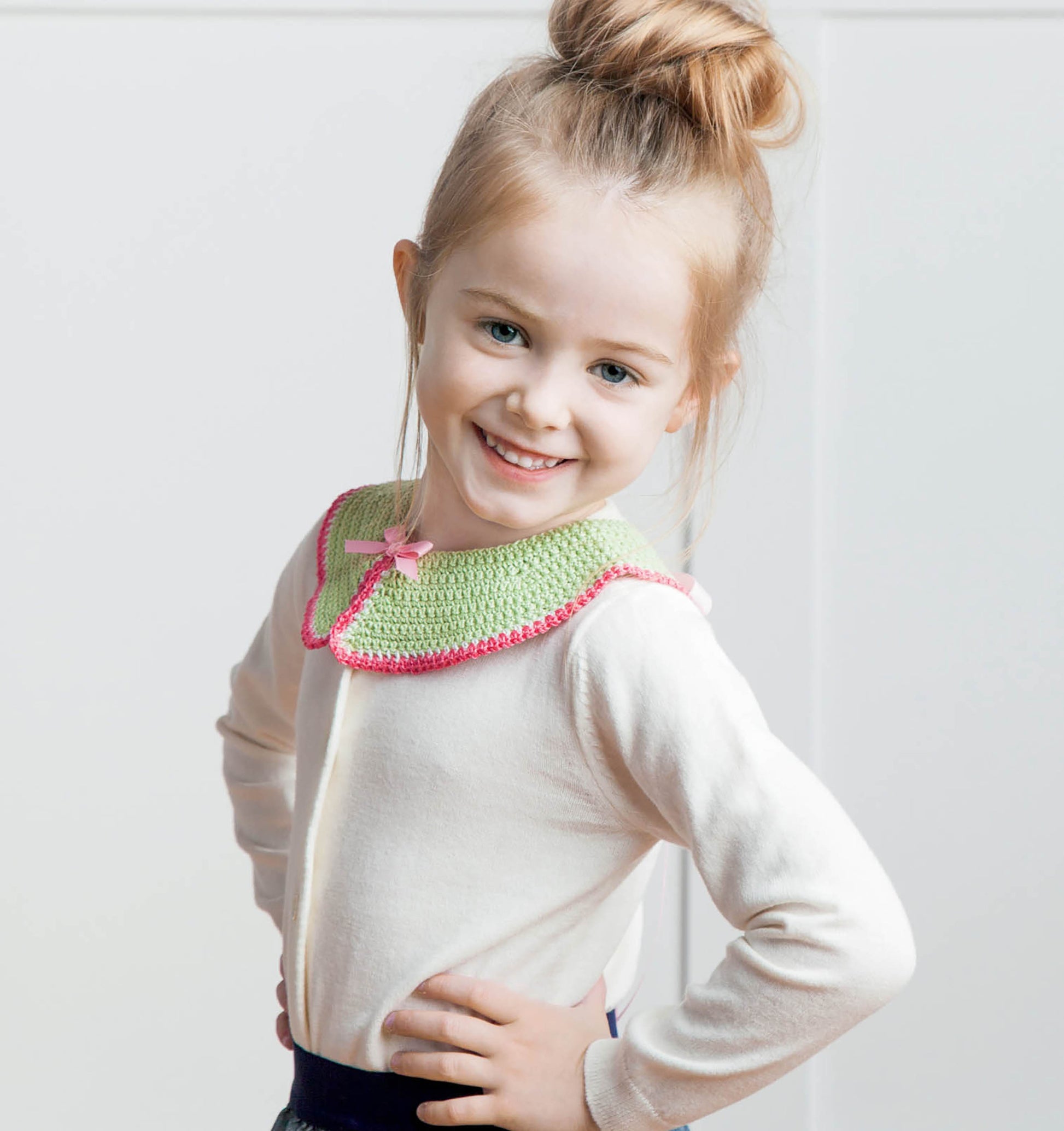 A smiling young girl with light brown hair in a bun models a white top and a green crochet collar with pink trim and bow from Leisure Arts' Crochet Collars for Kids—perfect inspiration for those seeking cute, beginner-friendly crochet patterns.