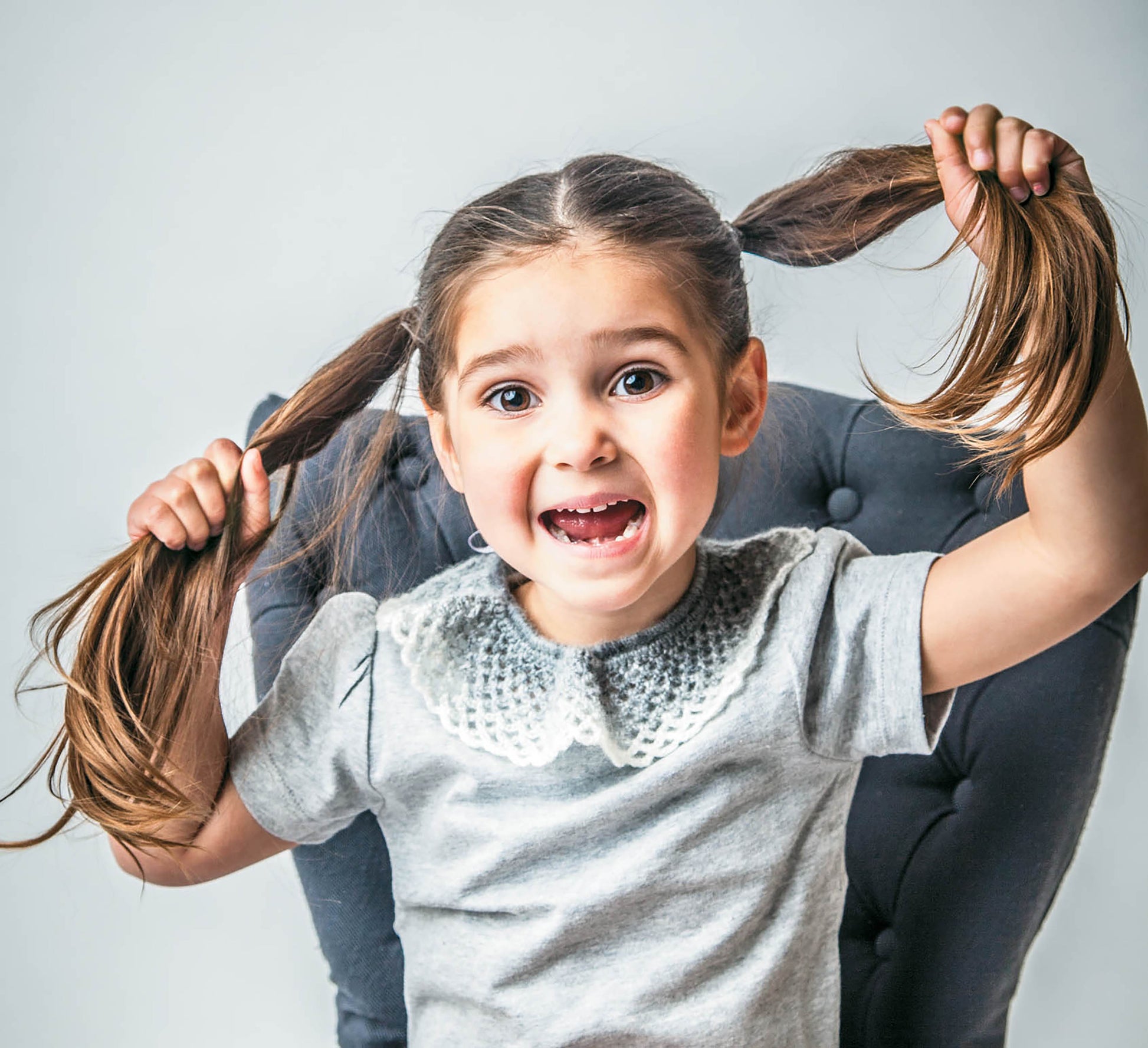 A young girl with brown pigtails sits on a gray chair, smiling and lifting her hair. She wears a light gray shirt accented by Leisure Arts' Crochet Collars for Kids, adding a charming handmade touch to her lively style.