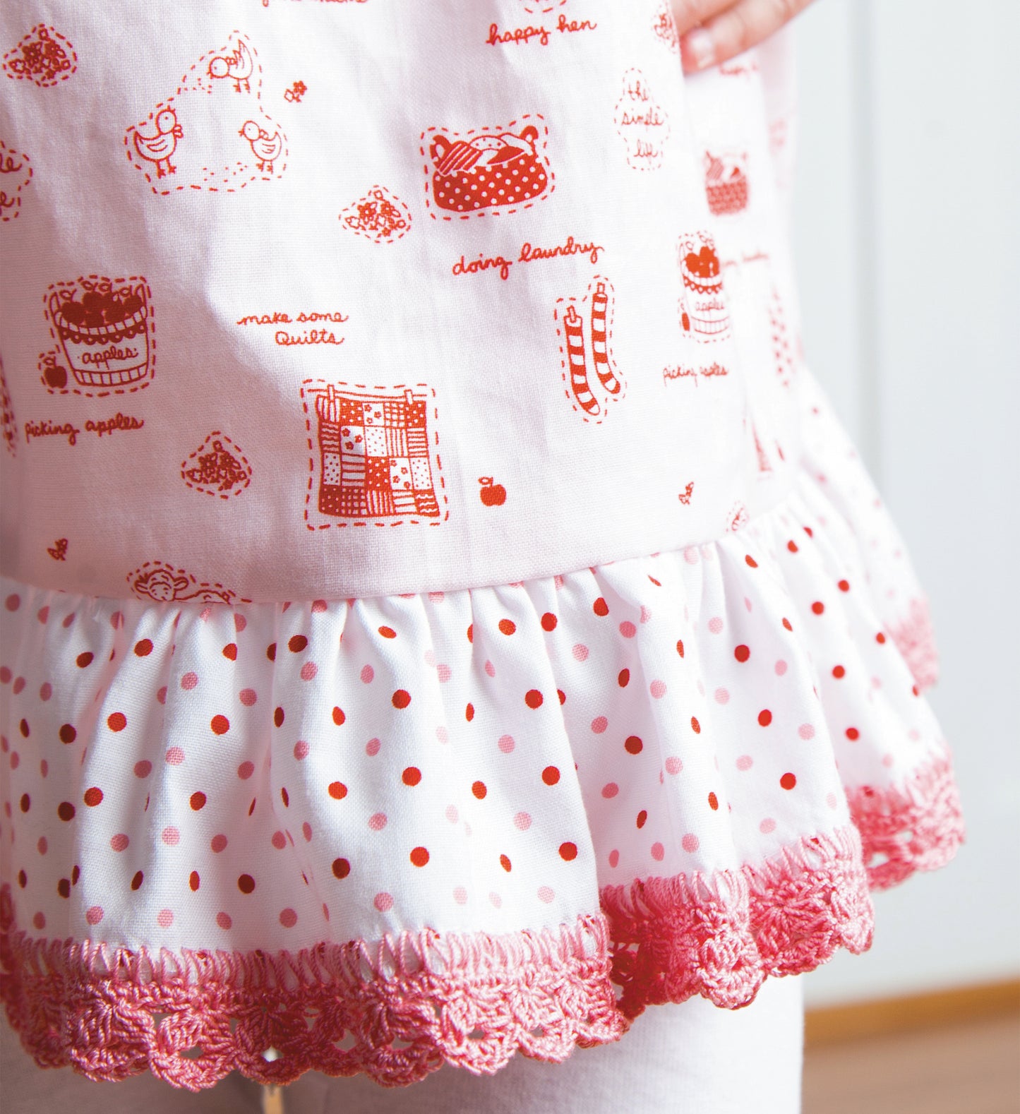 Close-up of a child's dress trimmed with pink beginner crochet edgings from Edgings for Kids by Leisure Arts, featuring pink and white fabric with red illustrations, text, a ruffled white polka-dot layer, and pink-trimmed hem.