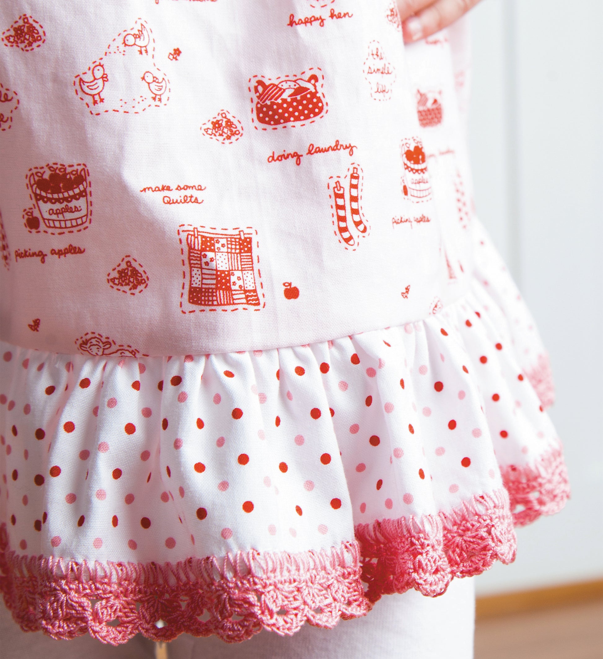 Close-up of a child's dress trimmed with pink beginner crochet edgings from Edgings for Kids by Leisure Arts, featuring pink and white fabric with red illustrations, text, a ruffled white polka-dot layer, and pink-trimmed hem.
