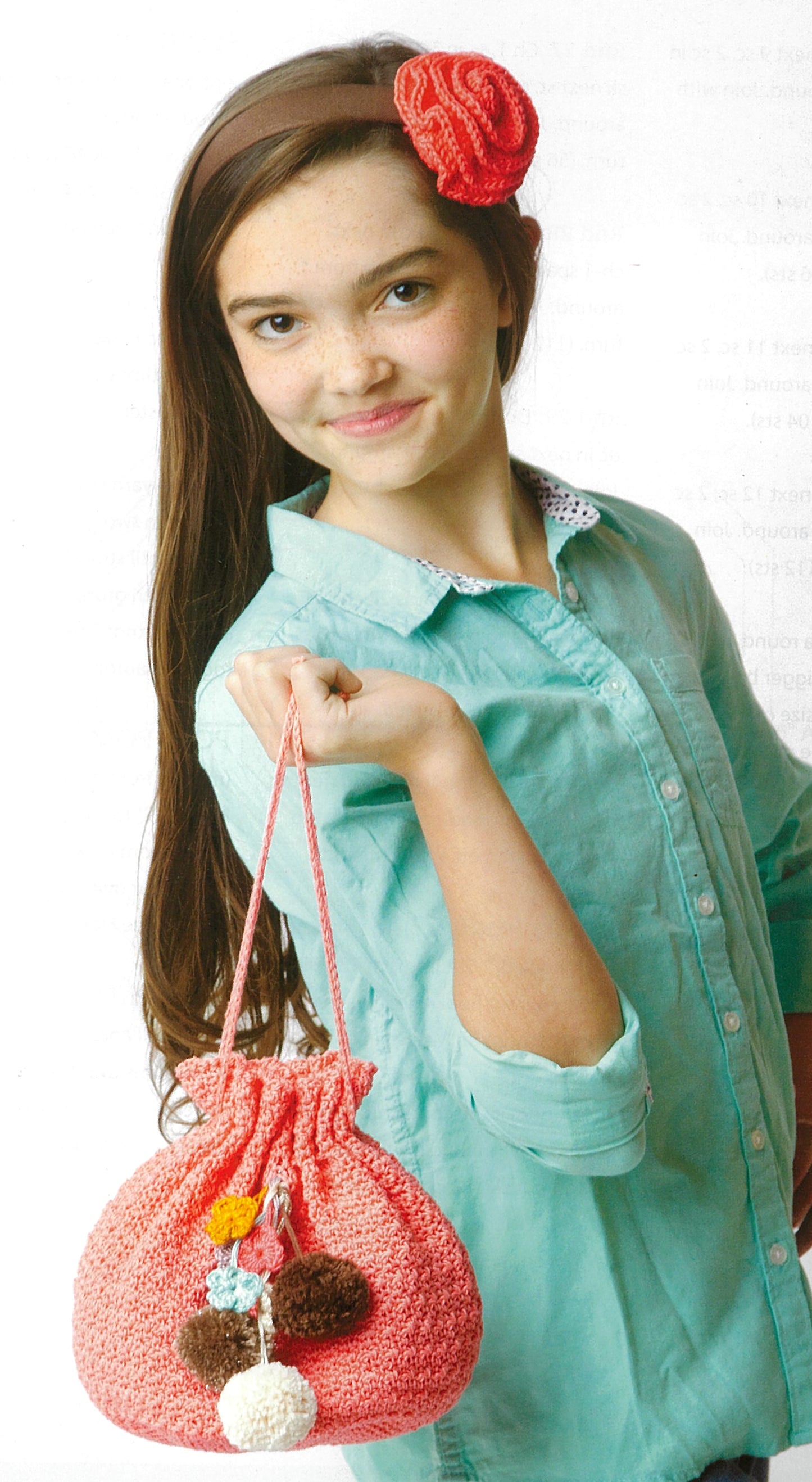 A girl with long brown hair, in a teal button-up and red hair accessory, smiles while holding a pink pom-pom drawstring bag—ideal for storing her Leisure Arts Learn to Crochet kit or special crochet hook.