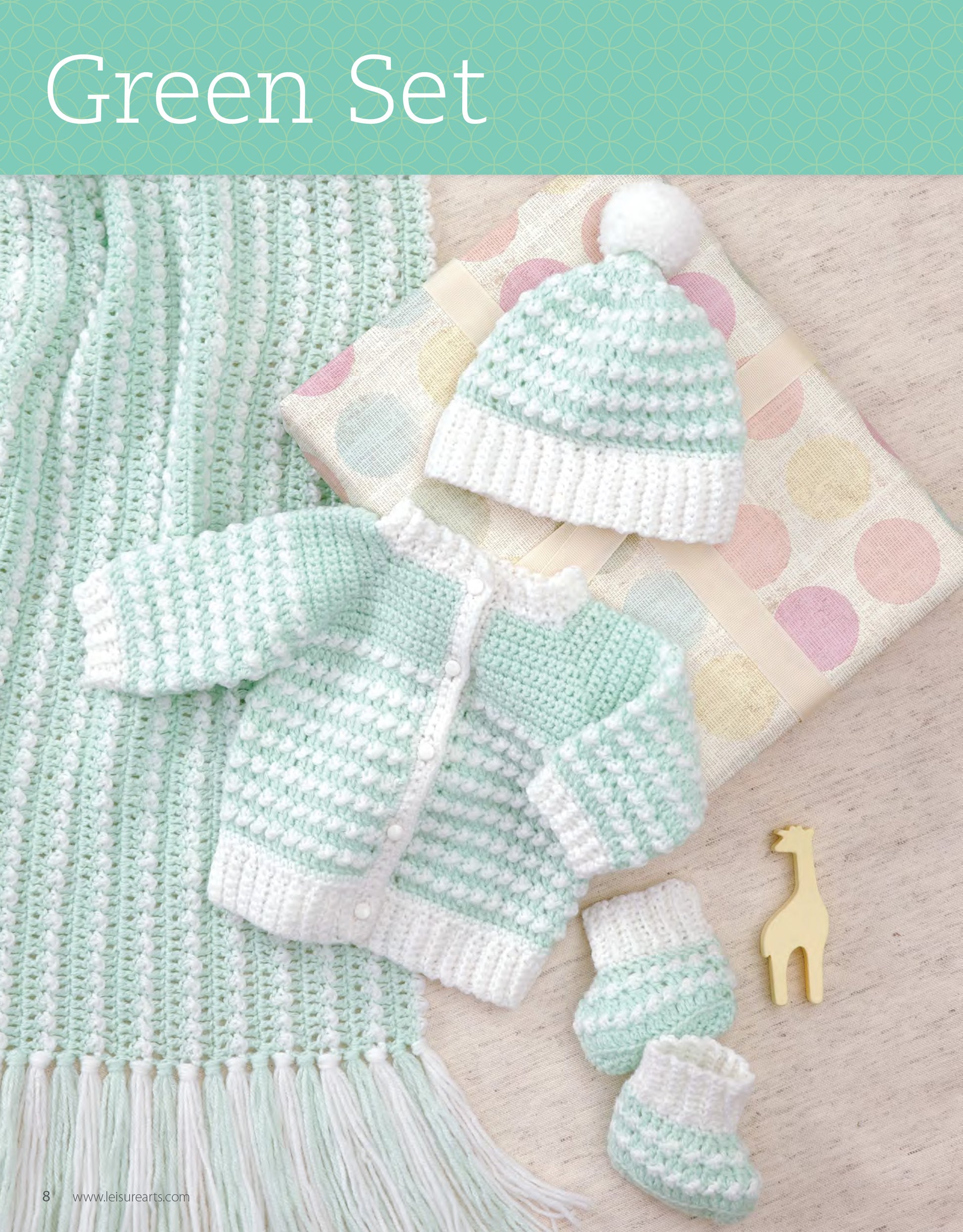 The My Very First Layette 2 by Leisure Arts is a mint green and white crocheted baby set with hat, sweater, and booties, shown on a cream background with pastel polka dot cloth, yellow giraffe toy, and fringed blanket.