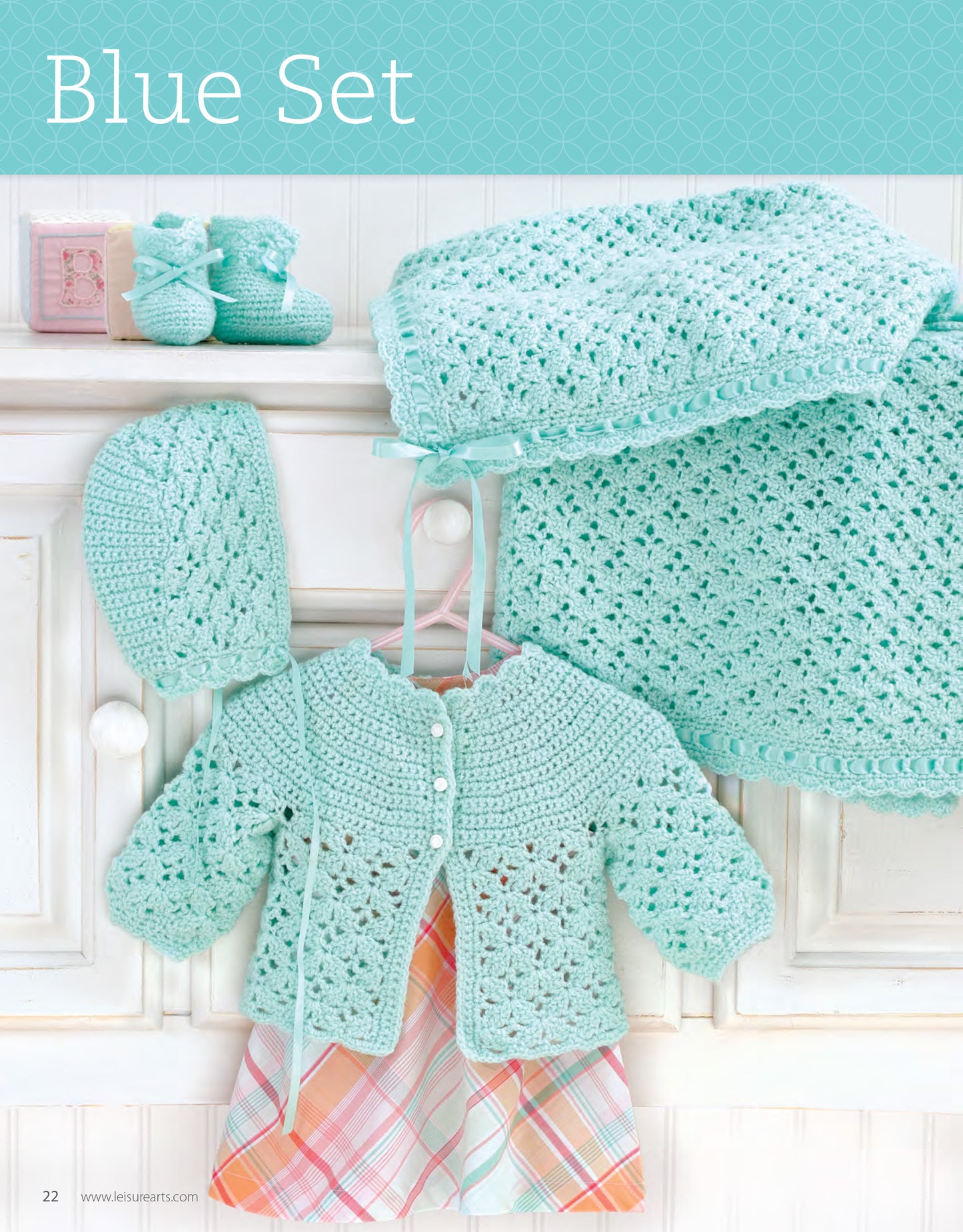 My Very First Layette 2 by Leisure Arts features a crocheted baby set in light blue with lacy patterns, displayed on a white cabinet. This easy crochet newborn pattern includes a cardigan, bonnet, booties, and blanket.