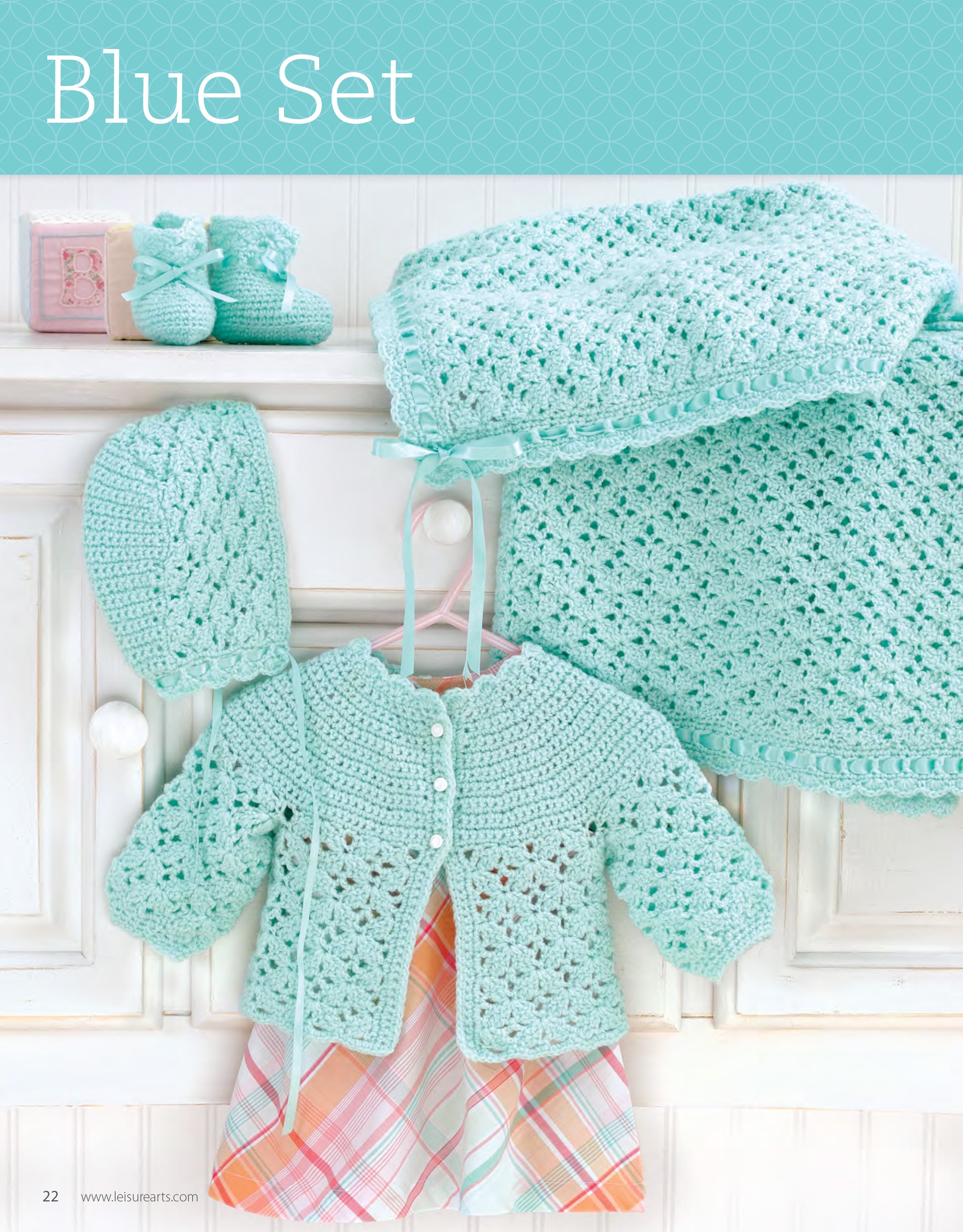 My Very First Layette 2 by Leisure Arts features a crocheted baby set in light blue with lacy patterns, displayed on a white cabinet. This easy crochet newborn pattern includes a cardigan, bonnet, booties, and blanket.