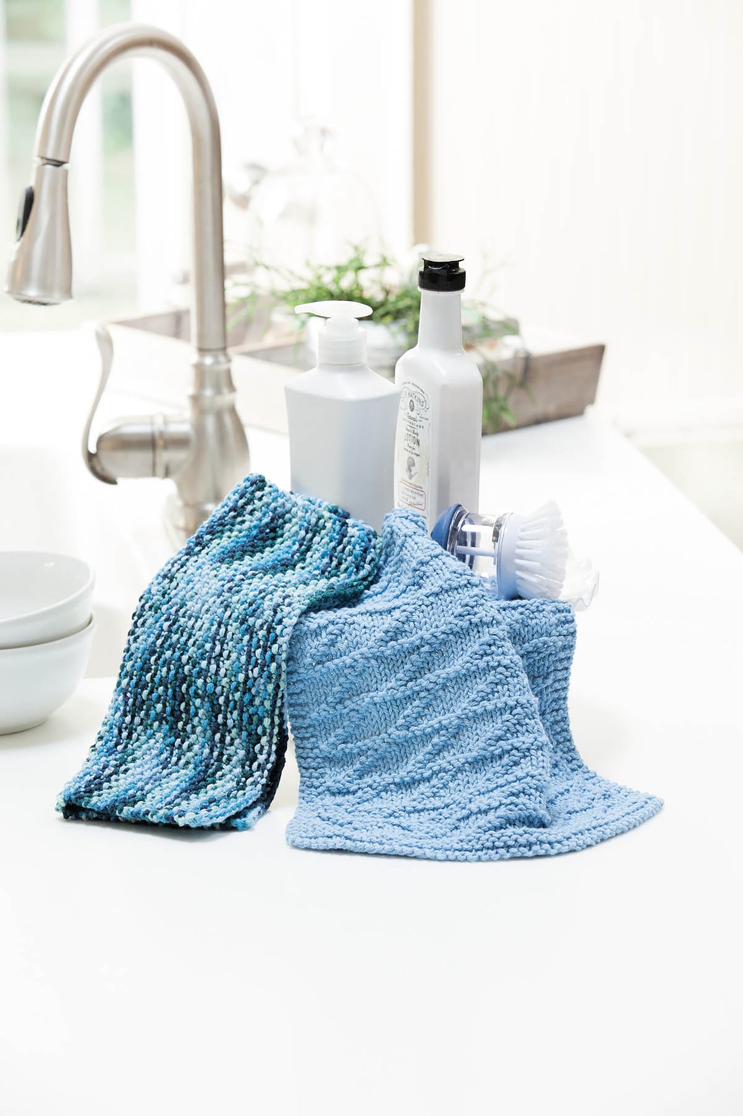 Two blue dishcloths made from Leisure Arts’ "10-20-30 Minutes to Learn to Knit" digital download are displayed on a white kitchen counter near a soap dispenser and cleaning items, with a faucet and blurred background visible.