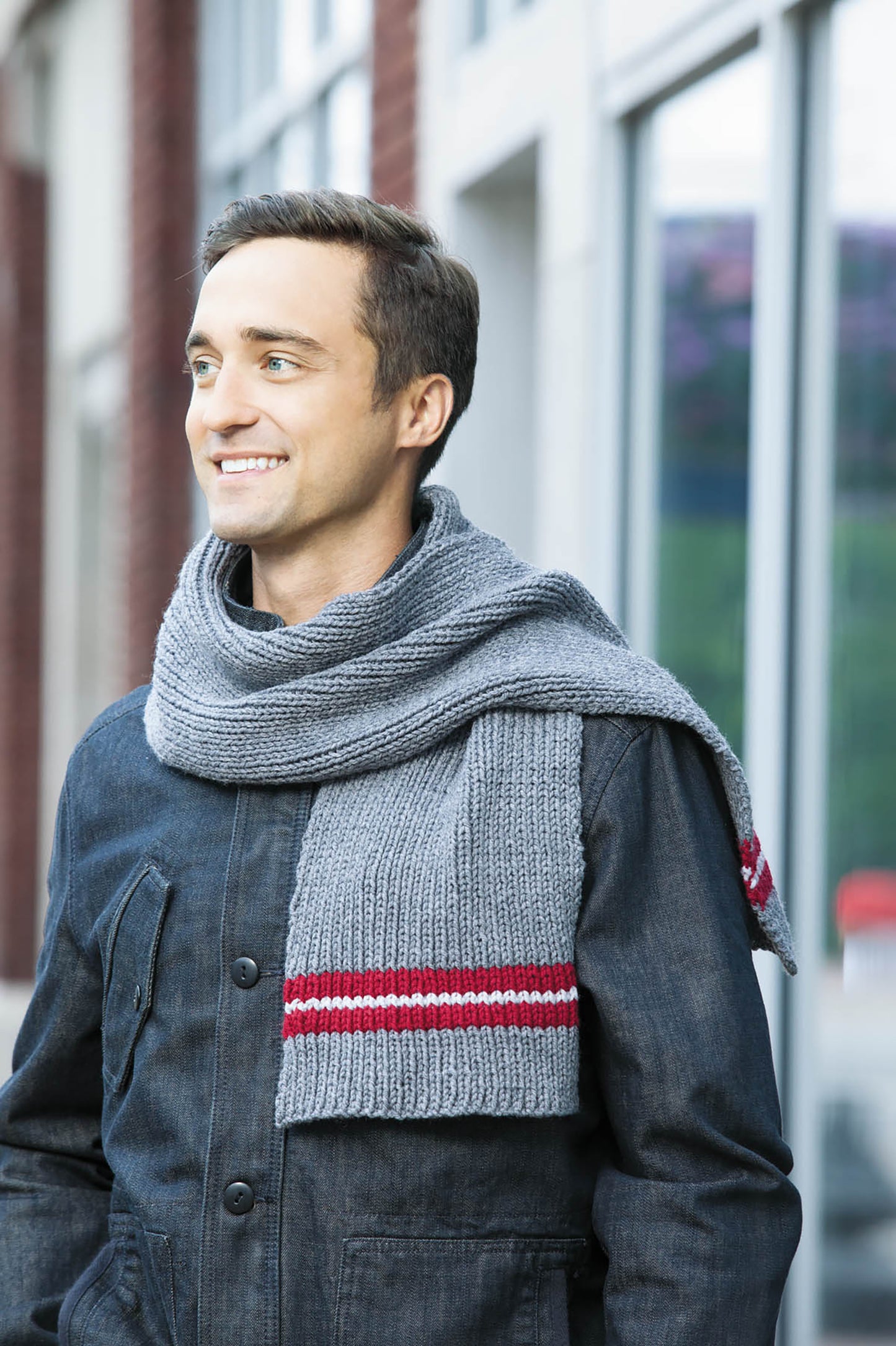 A man wears a gray scarf with red and white stripes, ideal for easy projects from *10-20-30 Minutes to Learn to Knit* by Leisure Arts, as he stands outdoors in front of a building, smiling and looking to the side.