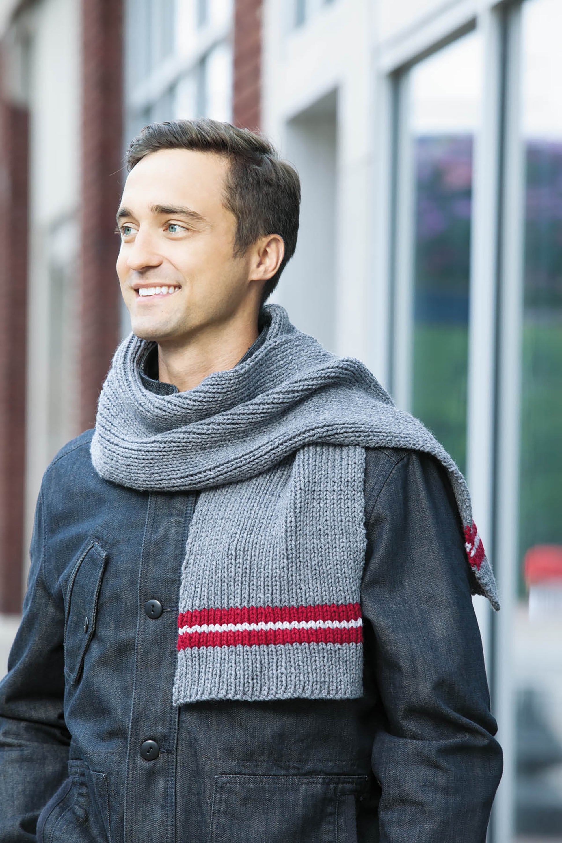 A man wears a gray scarf with red and white stripes, ideal for easy projects from *10-20-30 Minutes to Learn to Knit* by Leisure Arts, as he stands outdoors in front of a building, smiling and looking to the side.