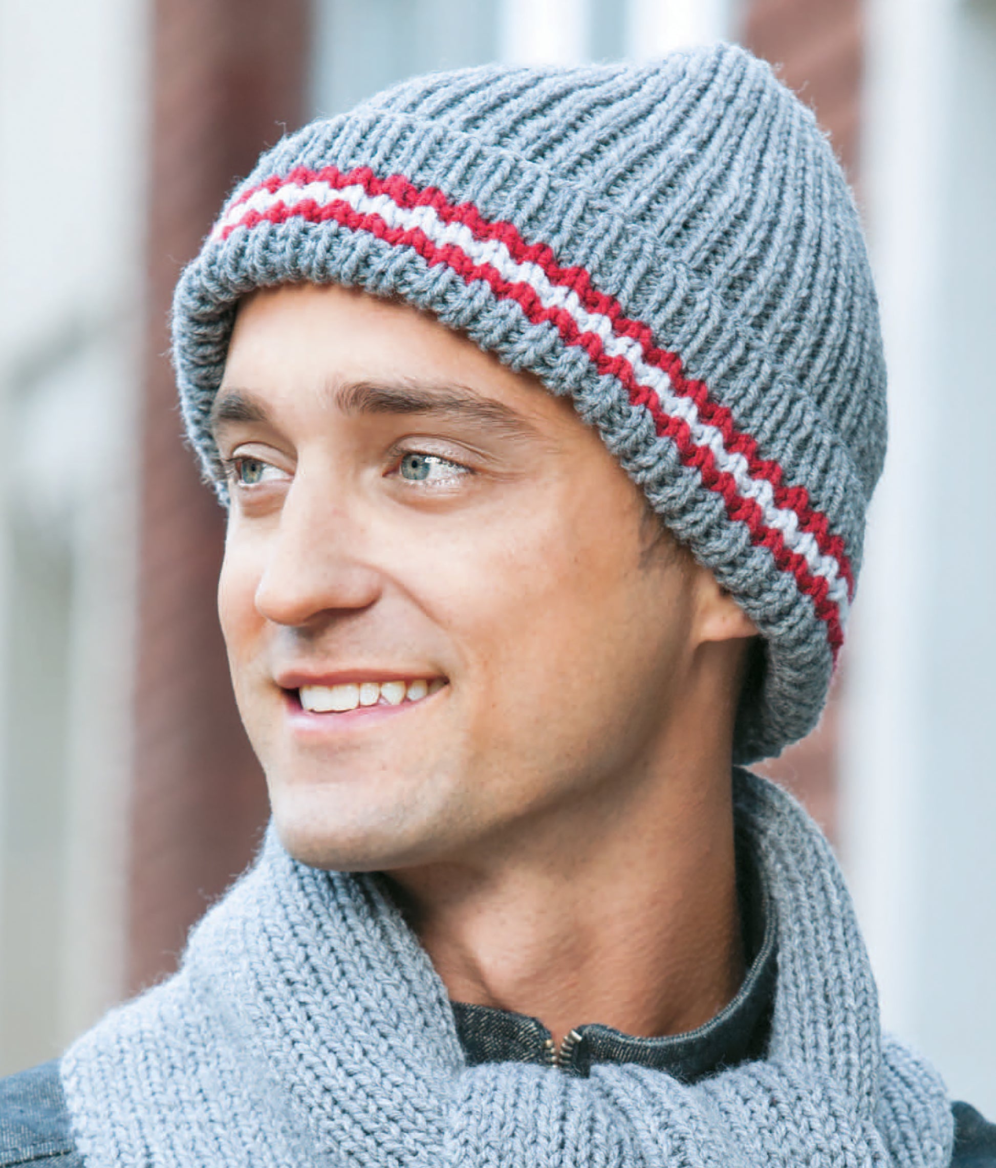 A man wearing a gray knit hat with red and white stripes, made from Leisure Arts' "10-20-30 Minutes to Learn to Knit," and a matching gray scarf, smiles and looks left in a blurred urban outdoor setting.