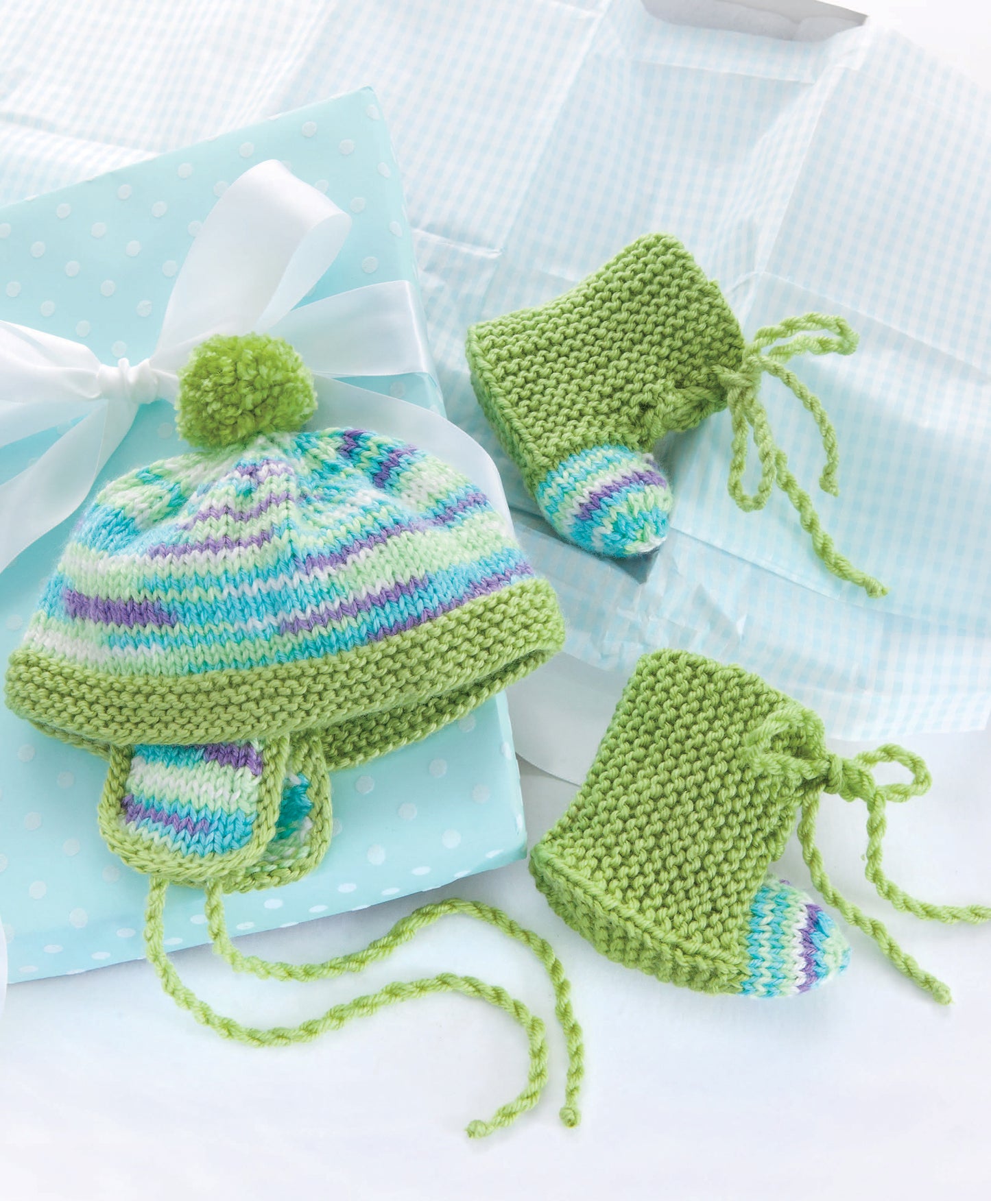 The Leisure Arts "10-20-30 Minutes to Learn to Knit" kit features a green, multicolored baby hat with pom-pom and matching booties, shown on light blue polka dot gift wrap tied with a white ribbon—ideal for easy knitting projects.