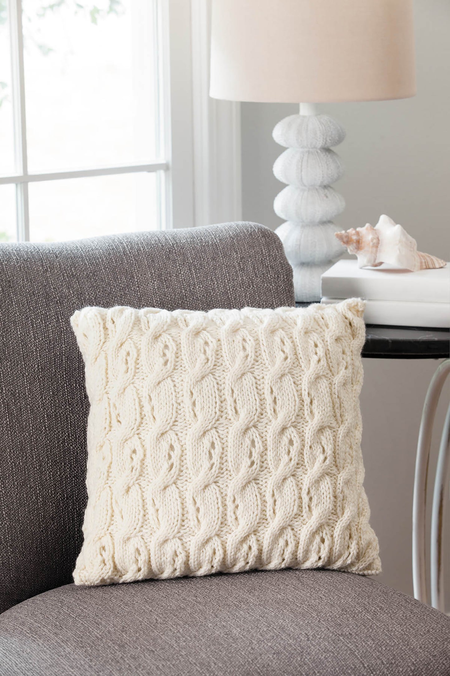 A cream knitted throw pillow, made using Leisure Arts’ “10-20-30 Minutes to Learn to Knit,” sits on a gray chair beside a table with a white lamp and seashell, while natural light streams in through the window.
