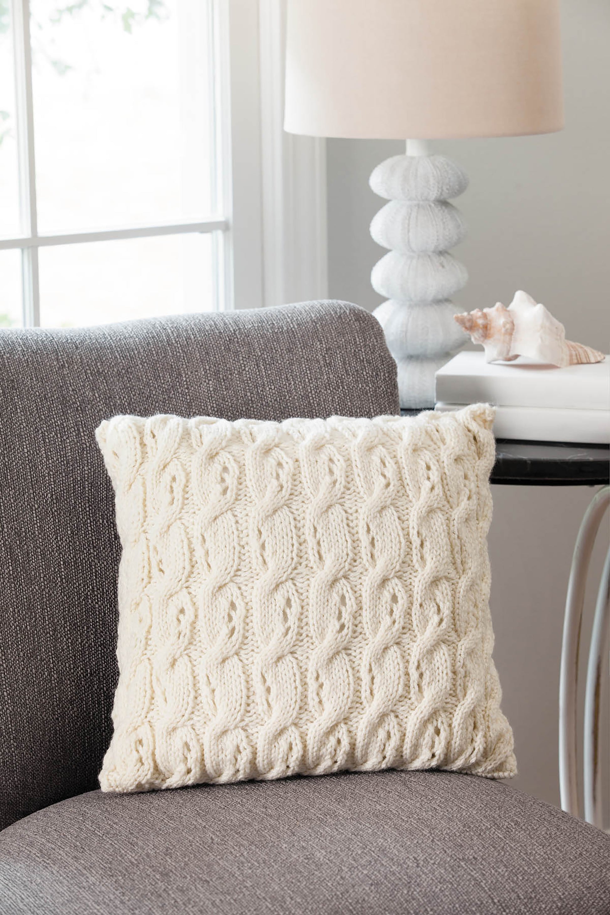 A cream knitted throw pillow, made using Leisure Arts’ “10-20-30 Minutes to Learn to Knit,” sits on a gray chair beside a table with a white lamp and seashell, while natural light streams in through the window.