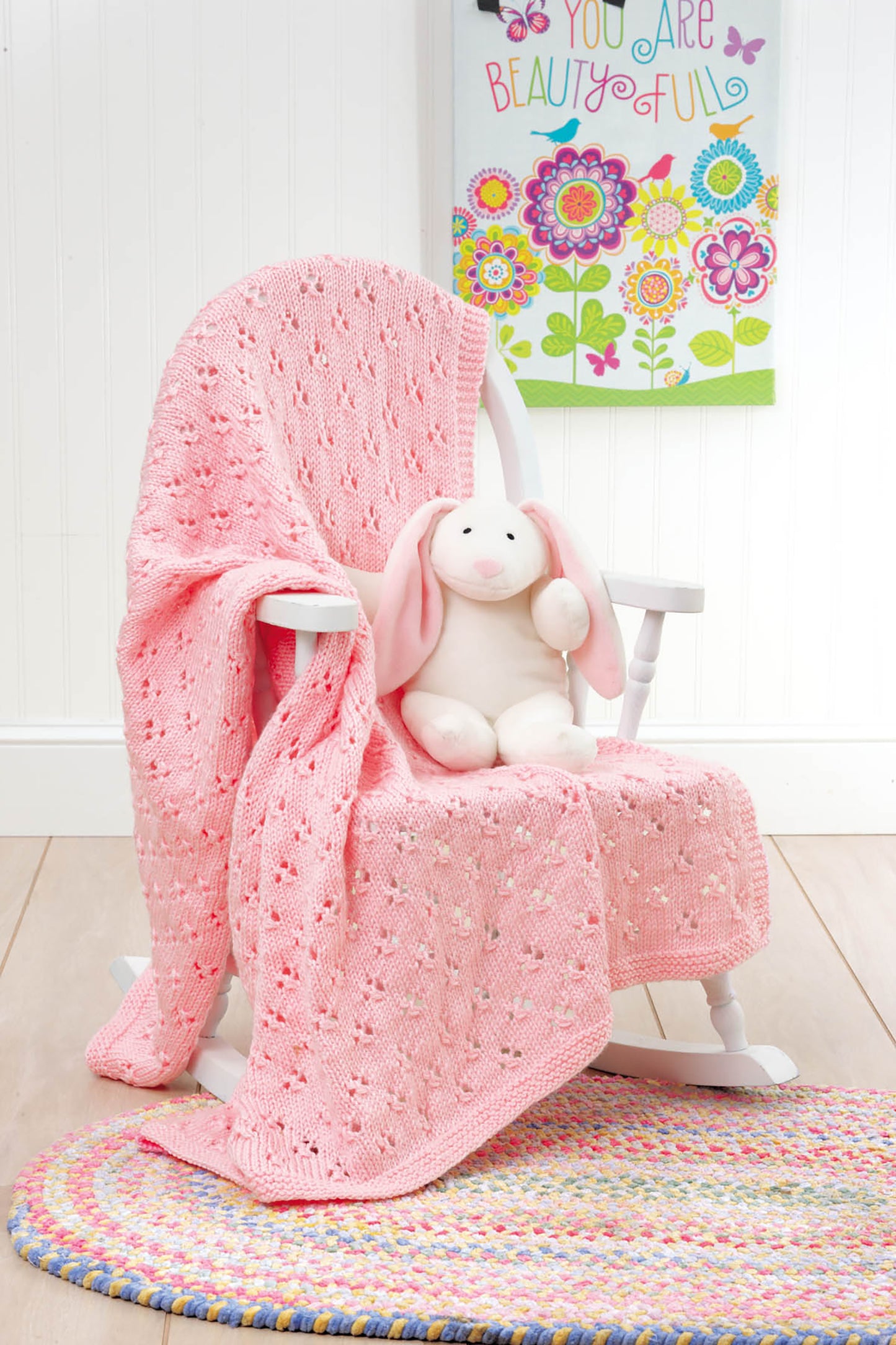 A white rocking chair with a pink blanket made from Leisure Arts' "10-20-30 Minutes to Learn to Knit" holds a plush bunny. A colorful rug is on the floor, and a bright floral "You Are Beautiful" sign decorates the wall behind.