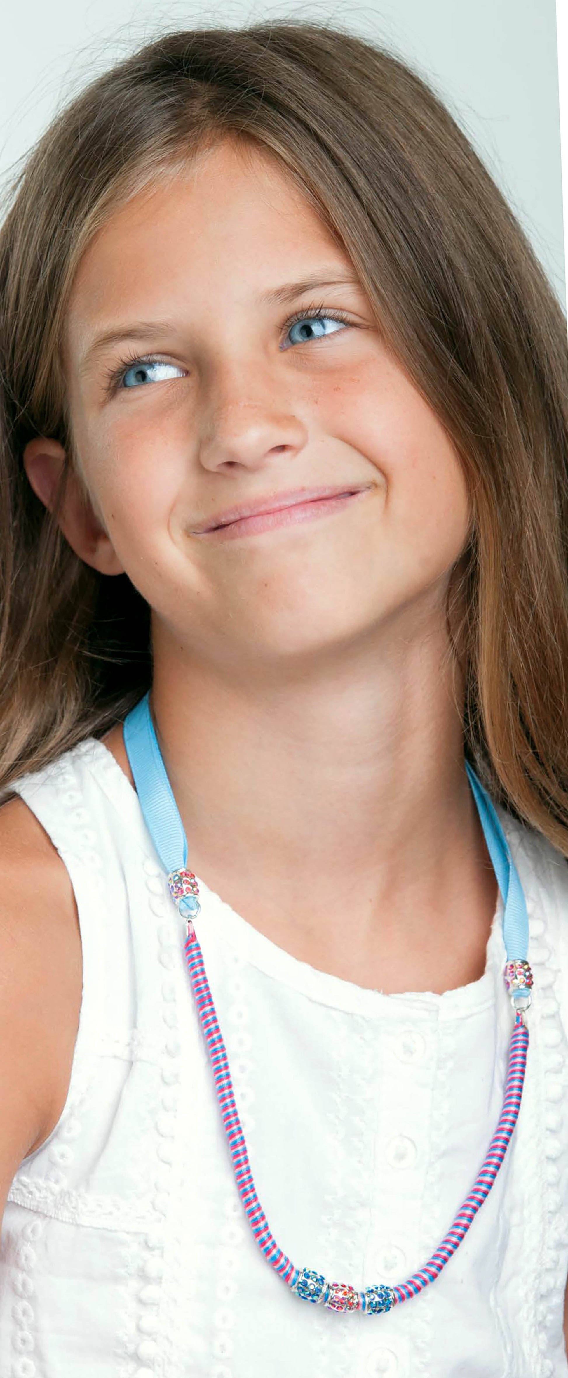 A young girl with long brown hair and blue eyes smiles softly, wearing a sleeveless white top and charming jewelry crafted from the “Amazing Jewelry Made Easy” kit by Leisure Arts, including a colorful beaded necklace with a blue ribbon and playful friendship bracelets that add a delightful touch to her look.