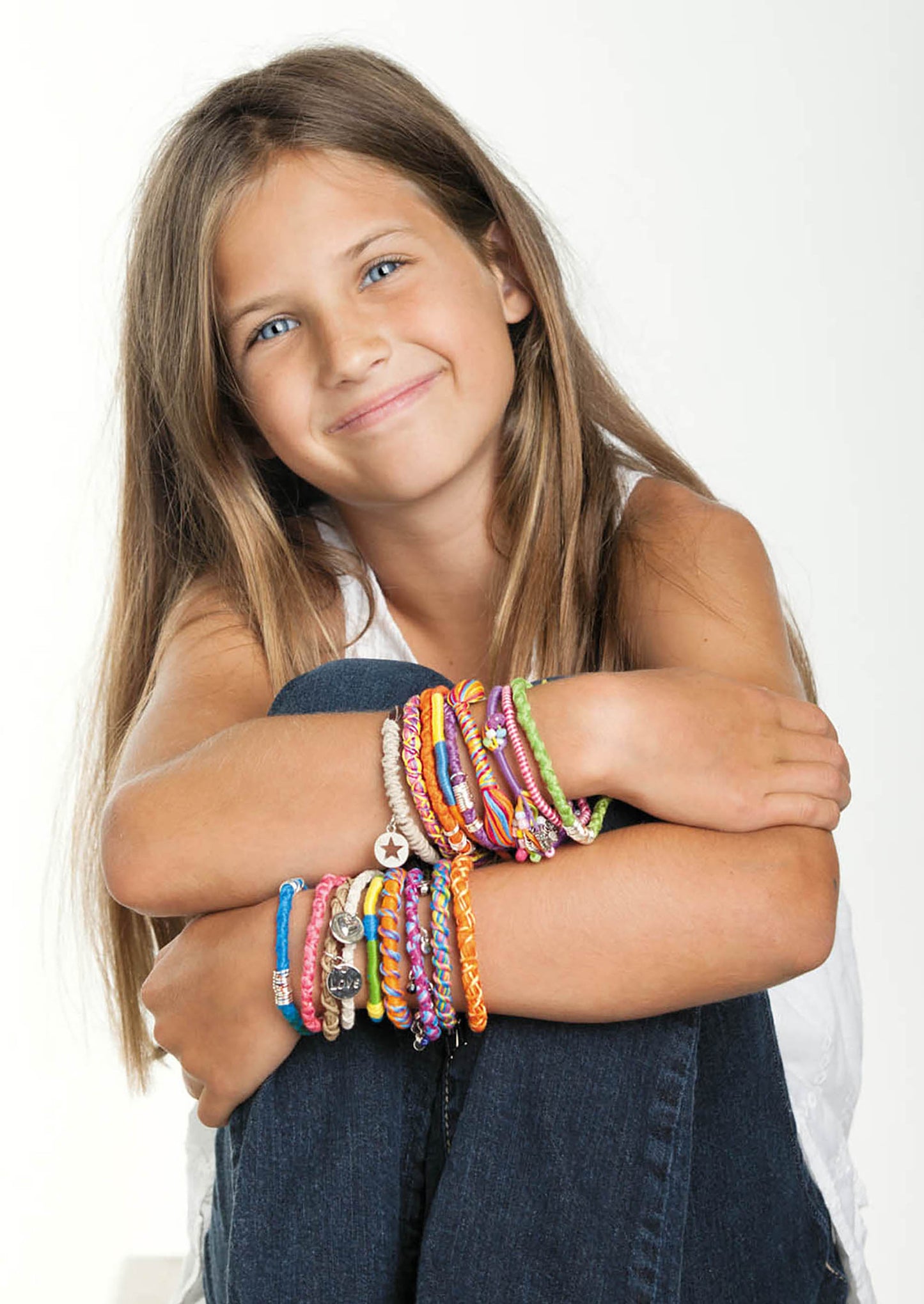A smiling girl with long brown hair, wearing a white shirt and blue jeans, sits with her arms crossed on her knees, proudly showcasing many colorful twisted accessories she created from the Amazing Jewelry Made Easy kit by Leisure Arts on both arms, against a plain light background.
