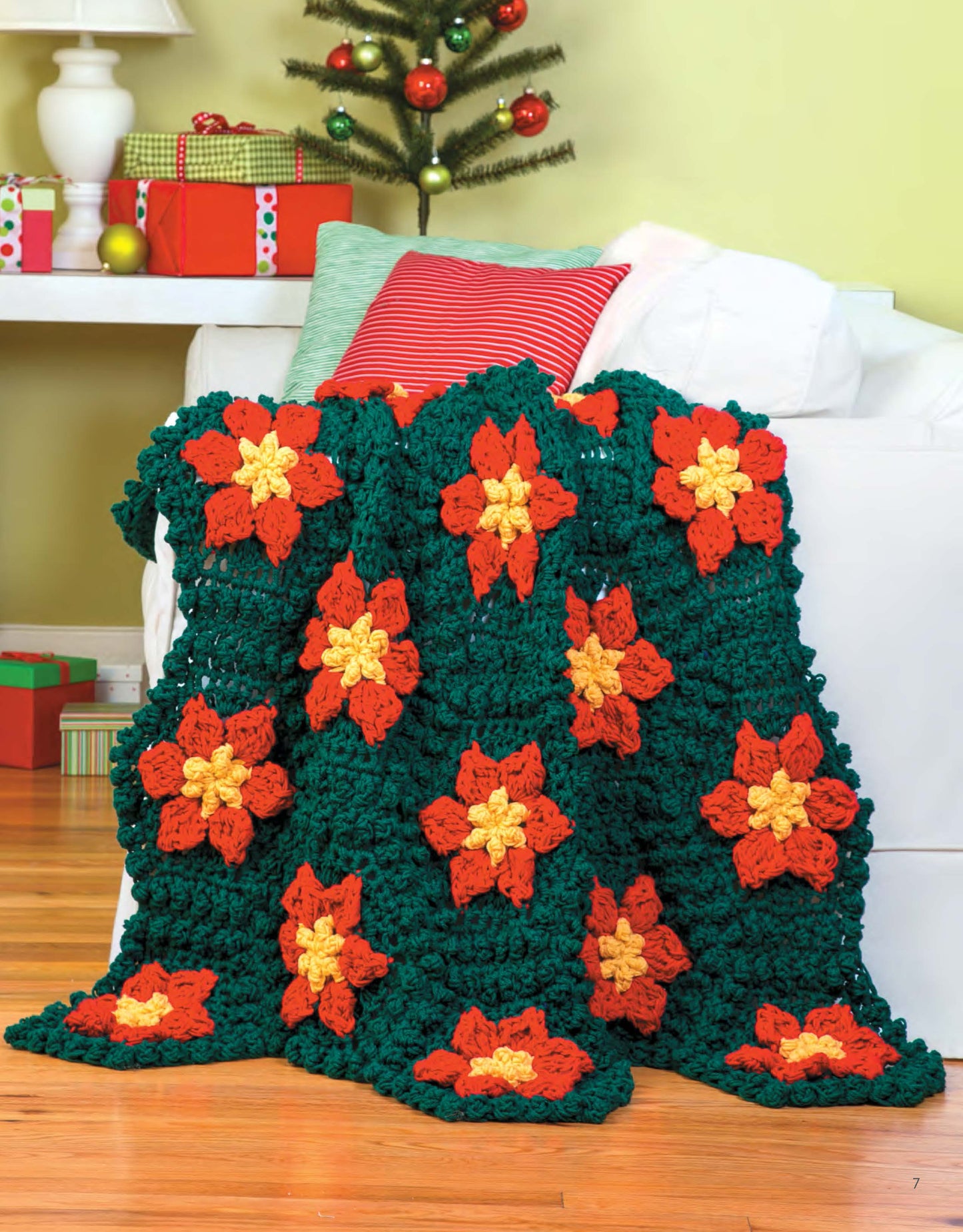 The Q-Hook Afghans for the Family Digital Download by Leisure Arts features a chunky crocheted blanket with red and yellow flower motifs on a green background, shown draped over a white couch near wrapped gifts and a small Christmas tree.