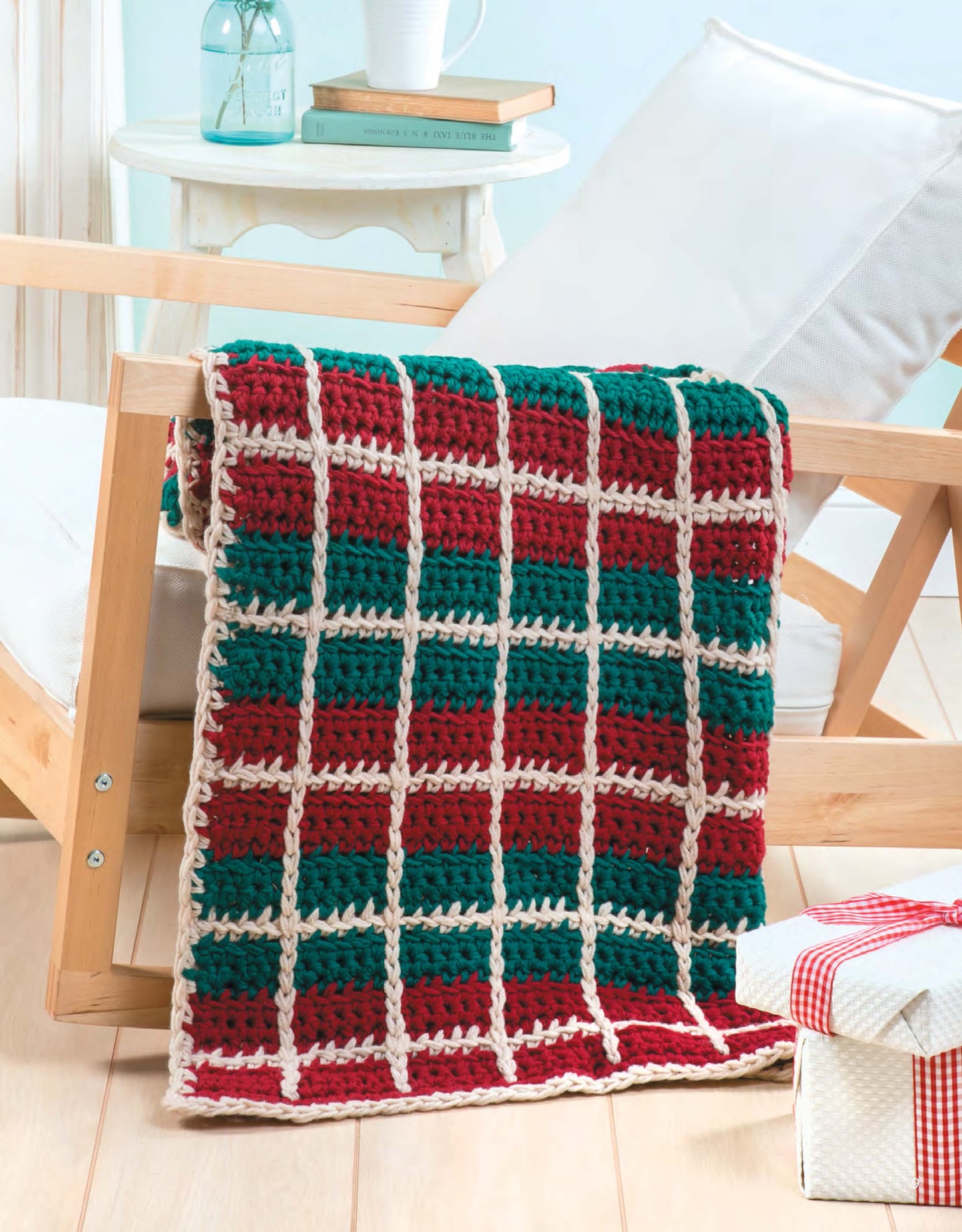 A Q-Hook Afghans for the Family crocheted blanket from Leisure Arts in red, green, and cream plaid drapes over a wooden chair. In the background is a white side table with books and a bottle; a wrapped gift sits on the floor nearby.