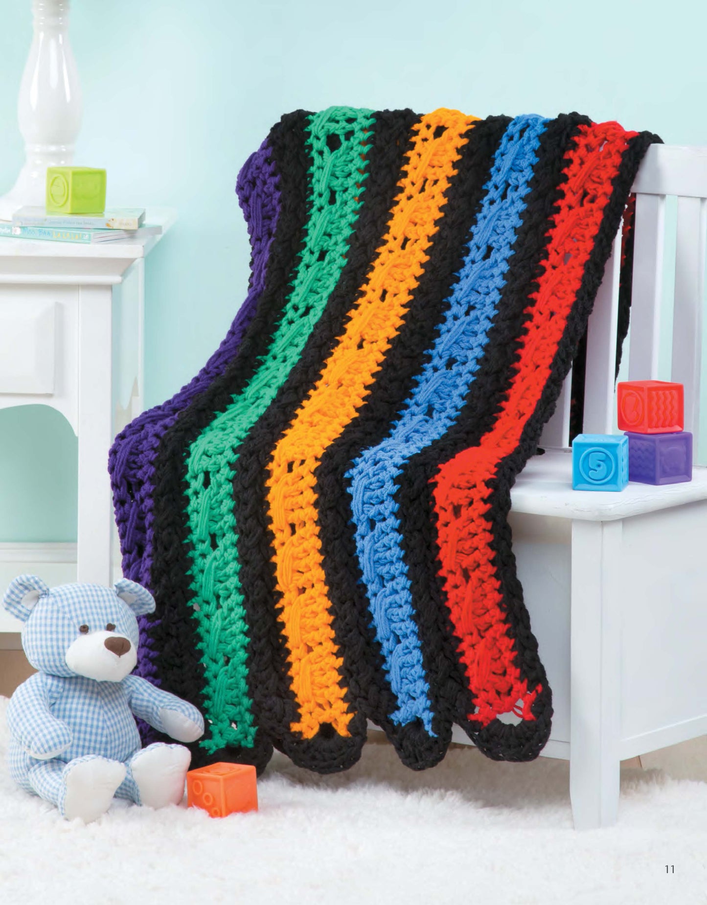The Q-Hook Afghans for the Family Digital Download by Leisure Arts features a vibrant crocheted blanket with black borders and bright stripes, shown draped over a white nightstand beside a blue teddy bear and colorful blocks.