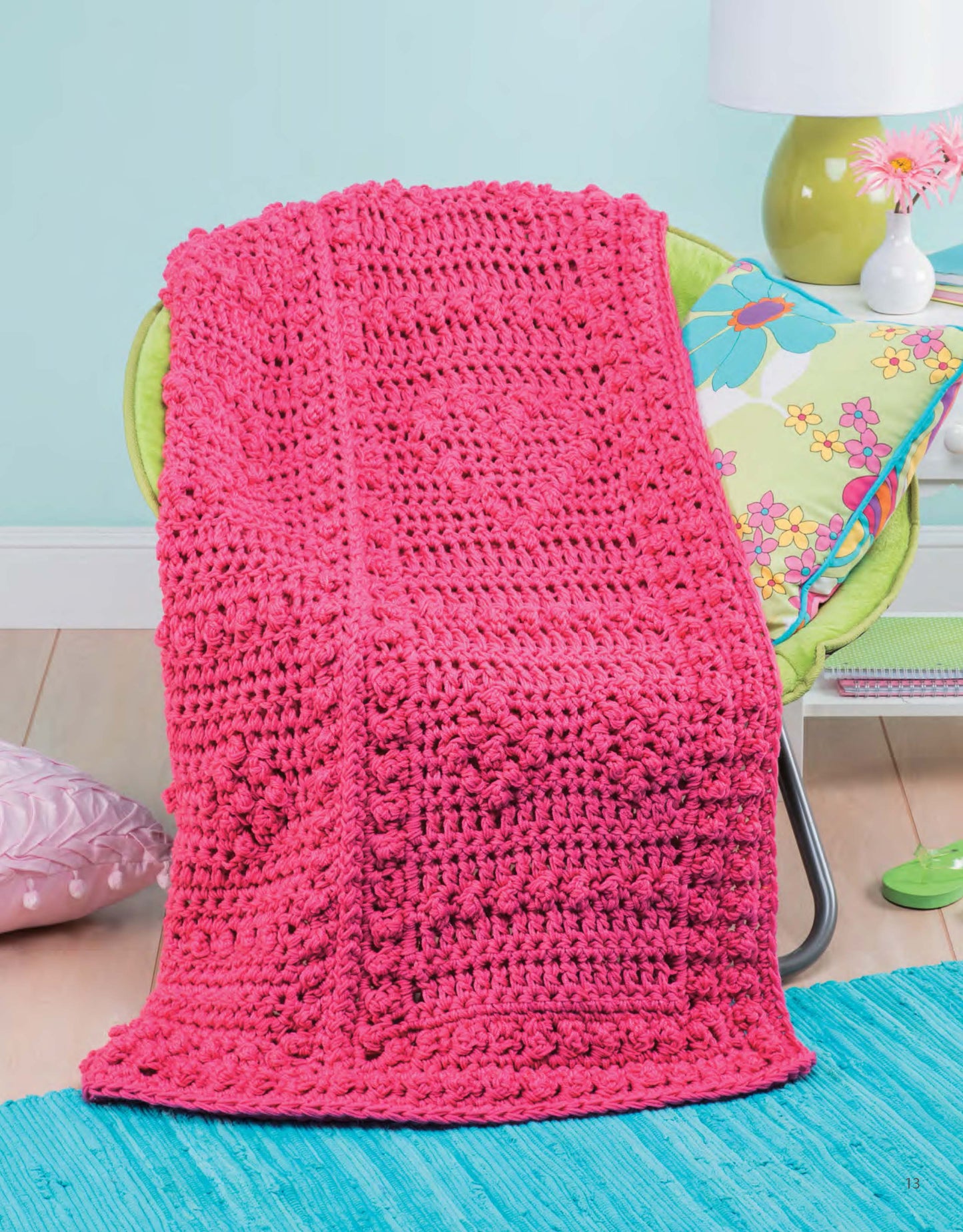 The Q-Hook Afghans for the Family Digital Download by Leisure Arts features a vibrant pink crocheted blanket with textured stitches, displayed over a green chair in a colorful room with a floral pillow and turquoise rug.