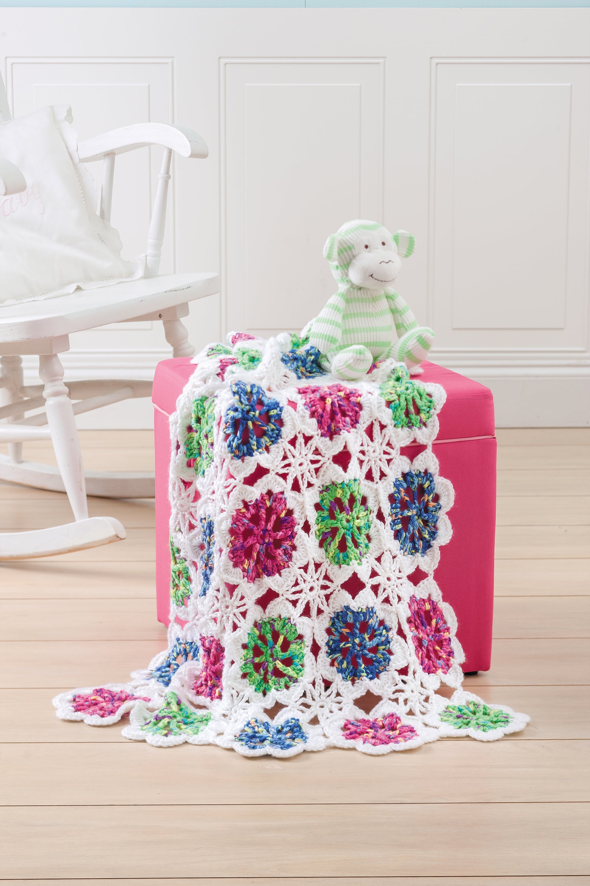 The Leisure Arts Crochet Refresher Digital Download features a vibrant crochet blanket with white borders and blue, pink, and green accents draped over a pink ottoman next to a green striped stuffed animal on a light wood floor.