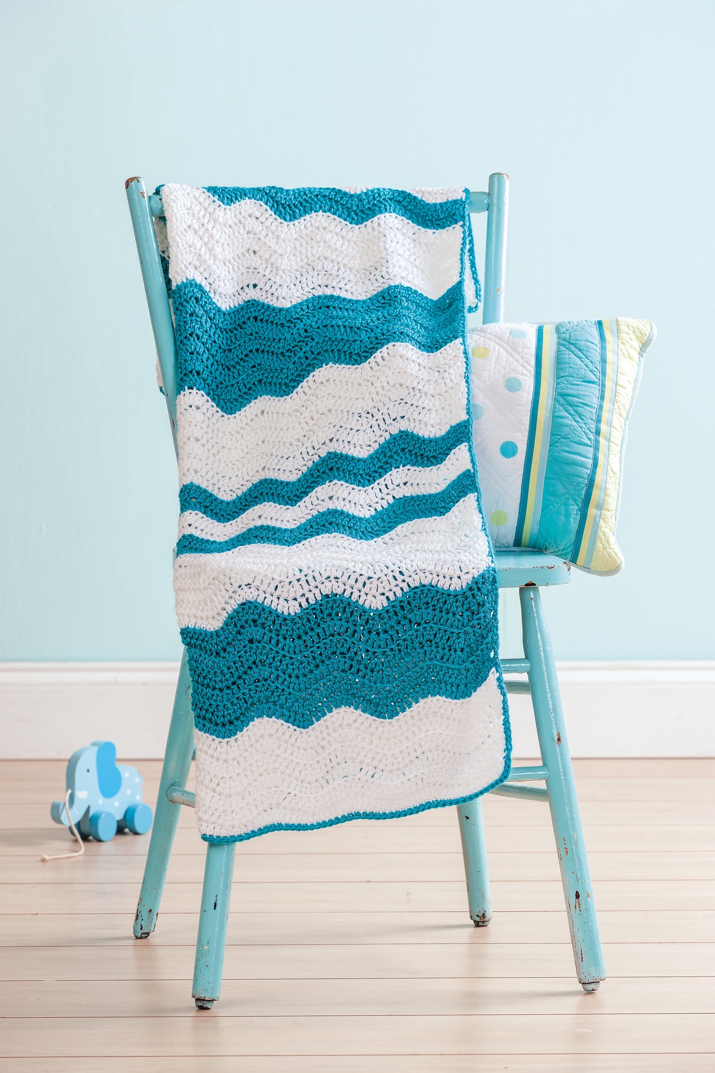The Crochet Refresher Digital Download by Leisure Arts features easy crochet patterns, shown in a blue and white striped blanket draped on a distressed blue chair with a patterned pillow and a blue elephant pull toy nearby.