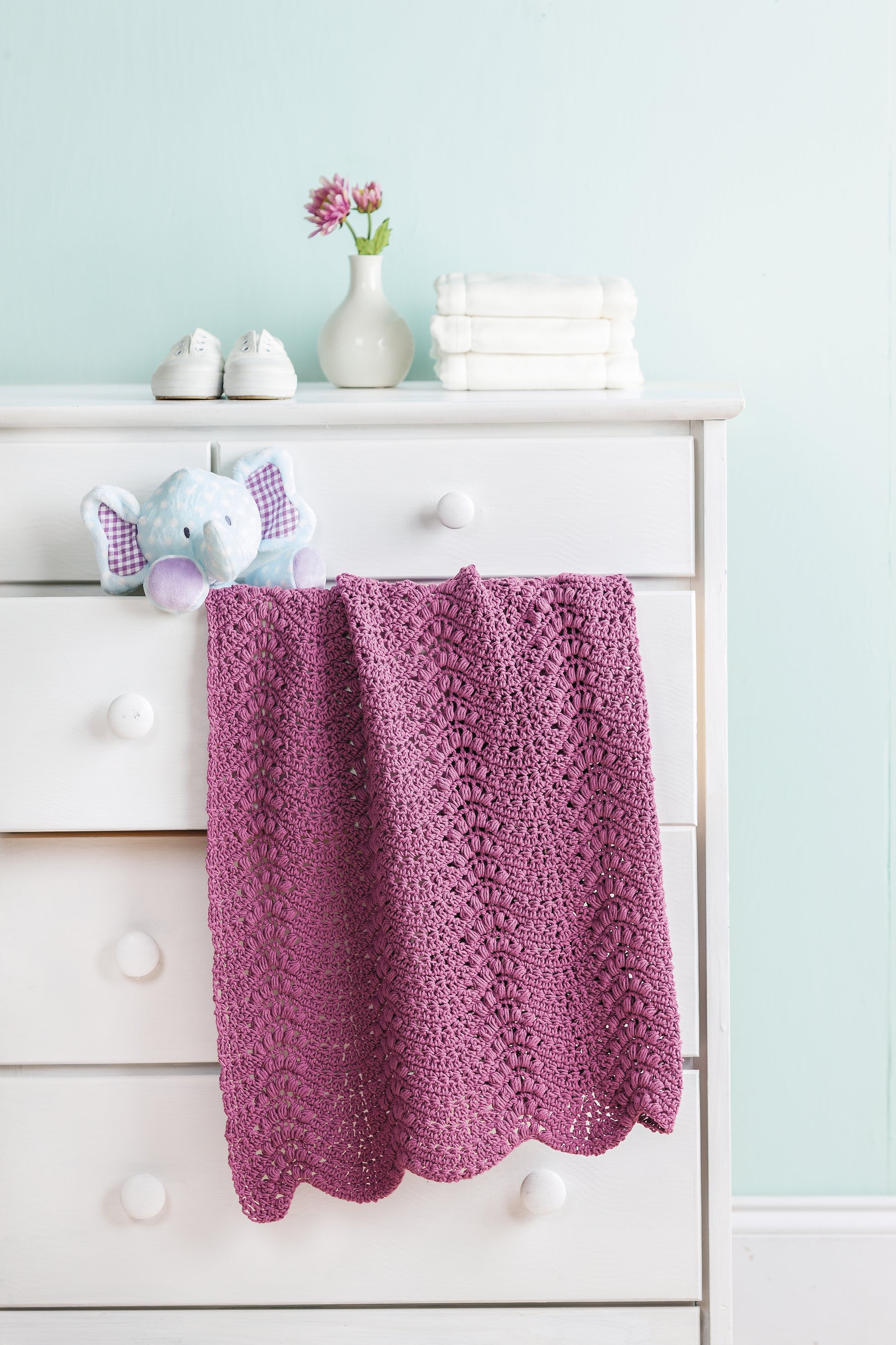 A white dresser with a Leisure Arts Crochet Refresher Digital Download-inspired purple crocheted blanket, blue stuffed elephant, white baby shoes, vase of pink flowers, and folded towels sits against a light blue wall.