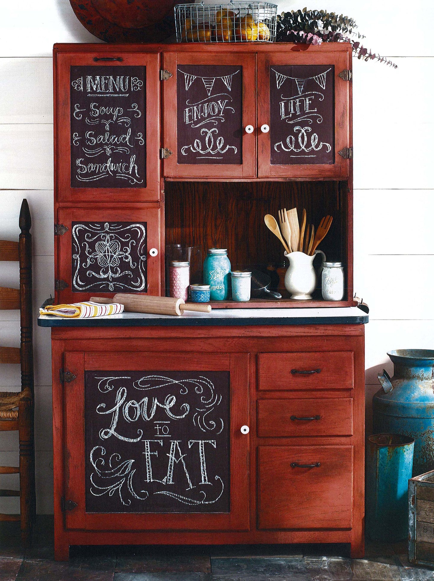 A copy of "A Beginner's Guide to Chalk Painting" by Leisure Arts is displayed on a rustic red kitchen hutch, surrounded by kitchen utensils, jars, and a rolling pin on the countertop. Chalkboard panels show handwritten menu messages.