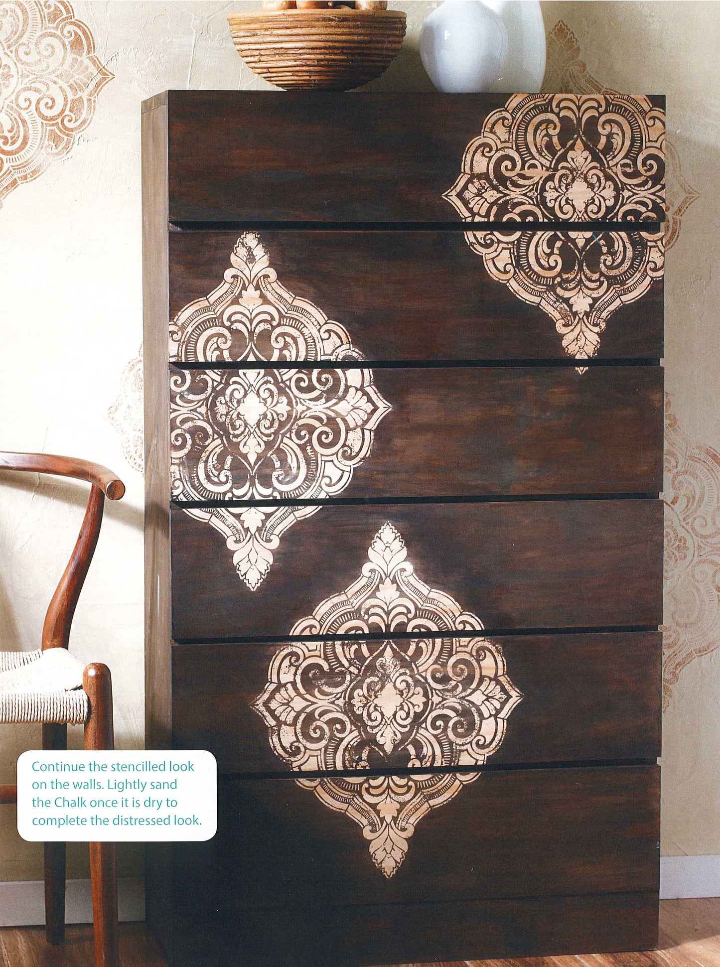 A dark wooden dresser features ornate, cream stenciled patterns inspired by "A Beginner's Guide to Chalk Painting" from Leisure Arts; matching designs extend onto the wall, creating a coordinated vintage look beside a wooden chair.