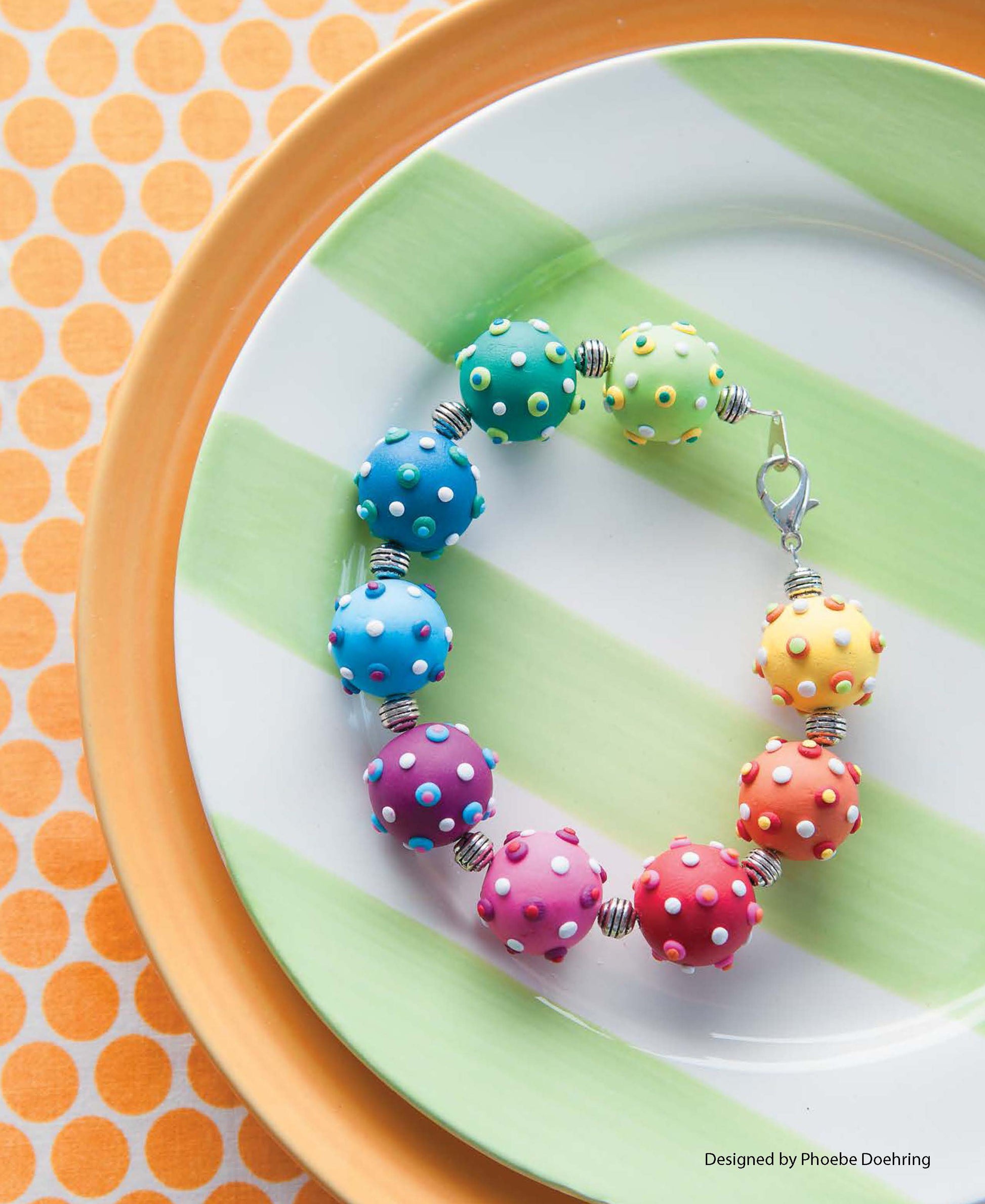 Leisure Arts Clay Jewelry featuring large, round, rainbow-colored beads with white polka dots is displayed on a white plate with green stripes atop an orange polka dot tablecloth.