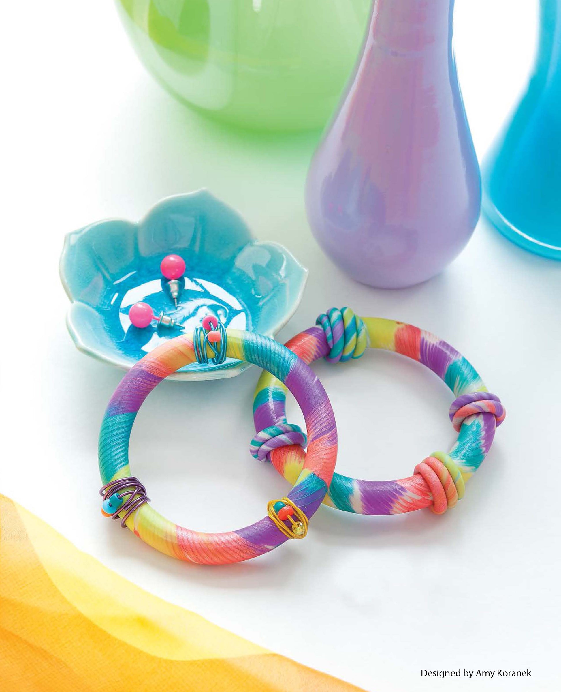 Leisure Arts Clay Jewelry: Two vibrant, tie-dye bangles with wire accents are shown on a white surface beside a blue dish with pink pins, pastel glass vases, and sheer yellow fabric.