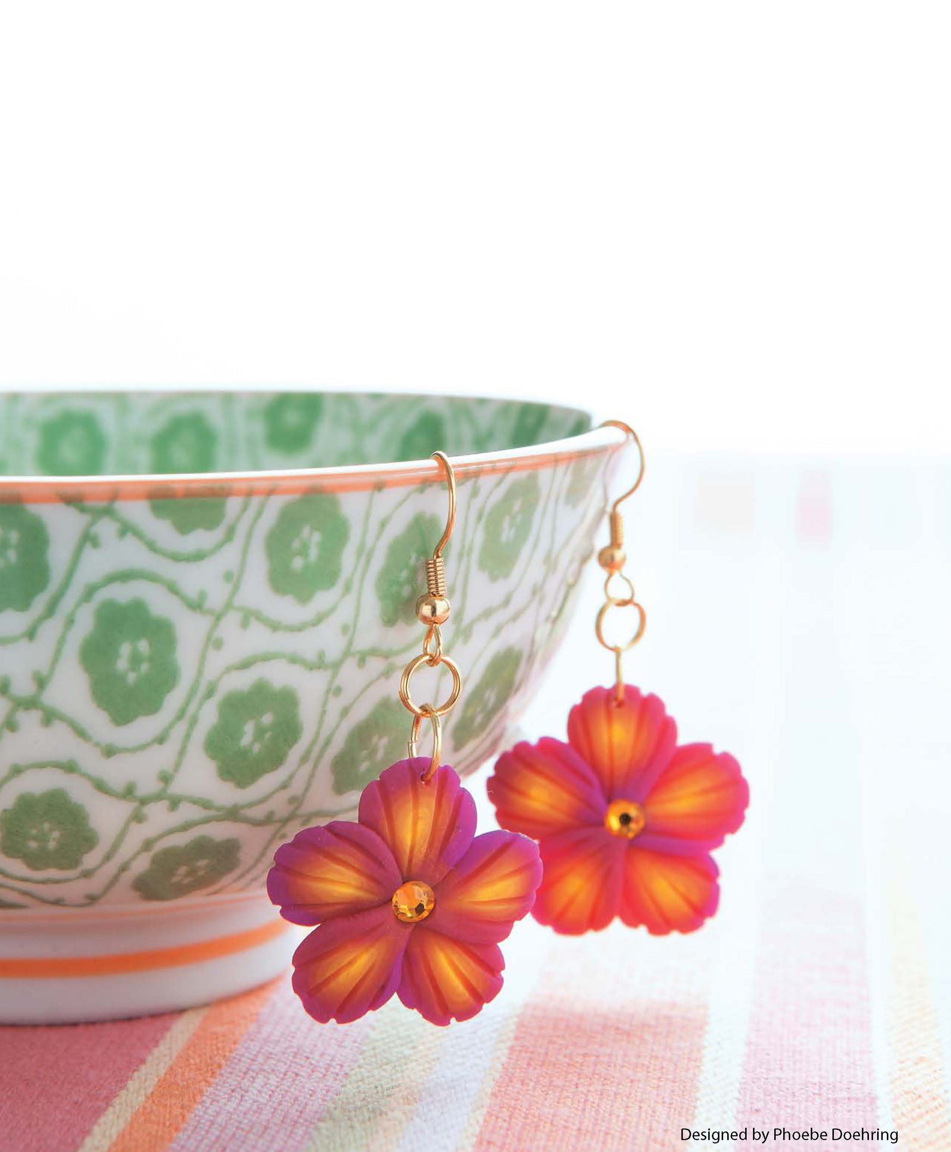A vibrant pair of Leisure Arts' Clay Jewelry flower earrings in orange and pink hangs from a patterned green-and-white bowl, set on a striped pastel surface.
