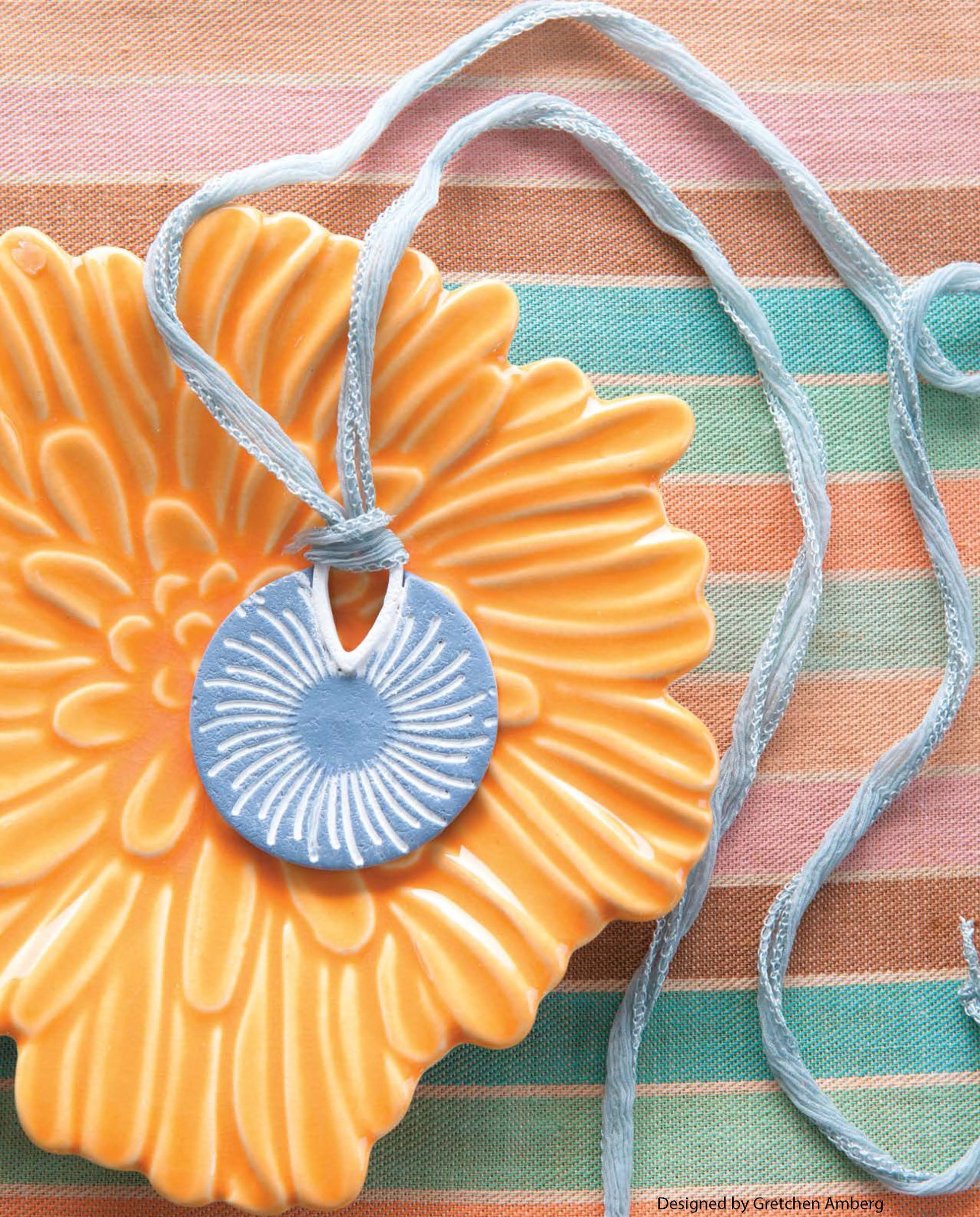 The Clay Jewelry by Leisure Arts features a round blue sunburst pendant on a light blue cord, displayed on an orange flower dish against pastel striped fabric.