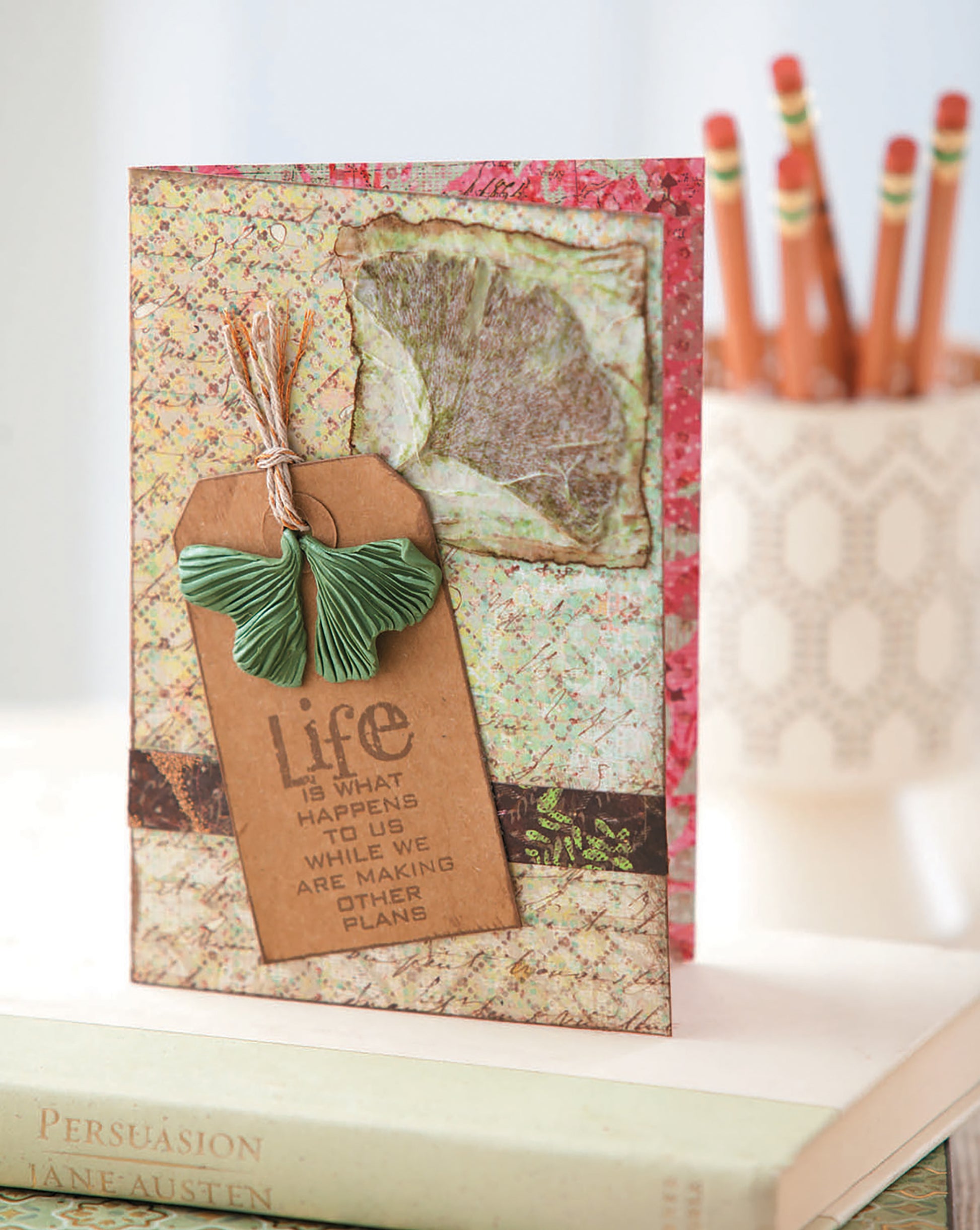 The book "Clay Creations for All" by Leisure Arts rests on a stack of books next to red pencils. A brown tag nearby reads, Life is what happens to us while we are making other plans.