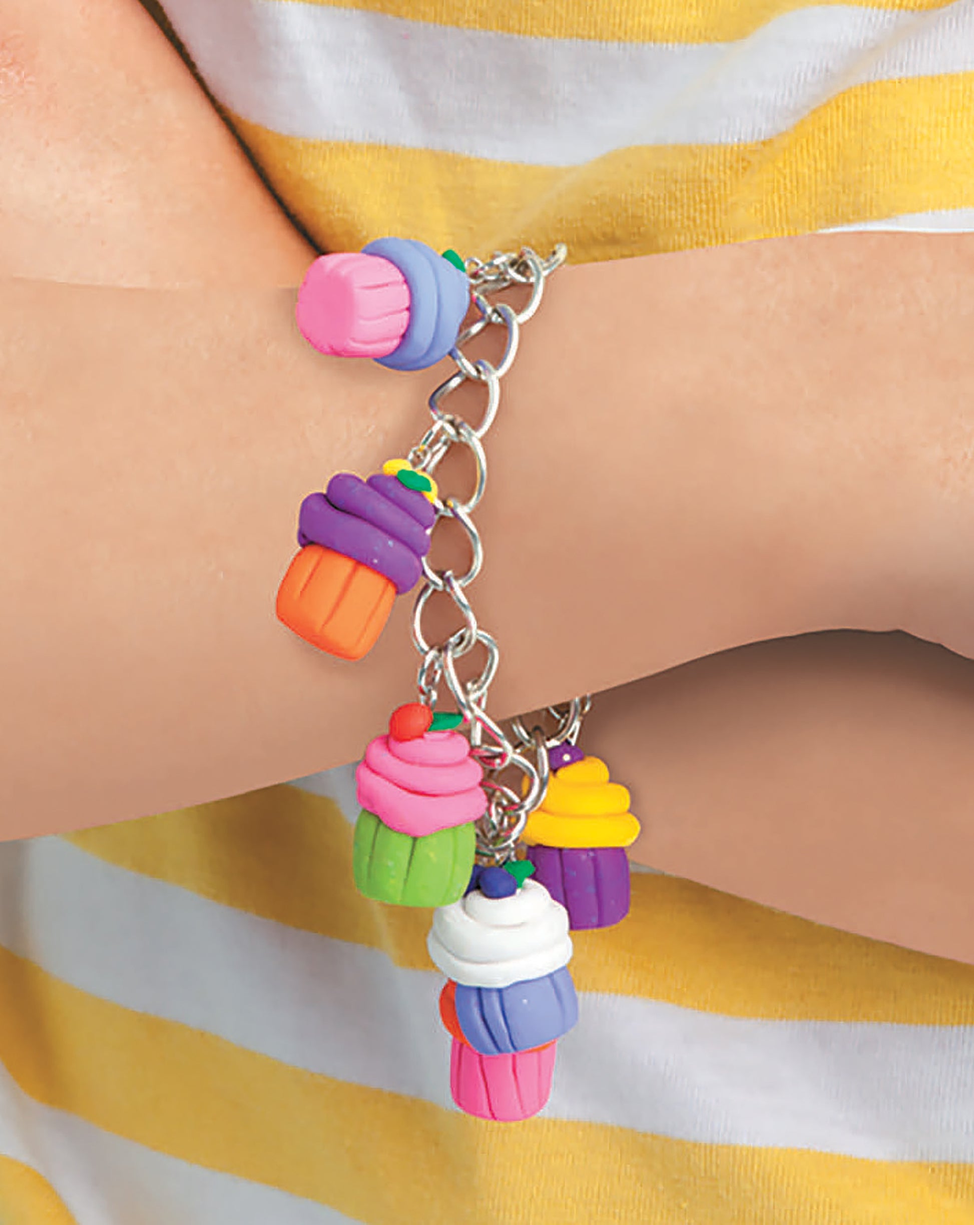 A person wearing the Clay Creations for All silver chain bracelet by Leisure Arts, featuring handmade polymer clay cupcake charms with unique decorations. Ideal for beginner crafters. The person wears a yellow and white striped shirt.