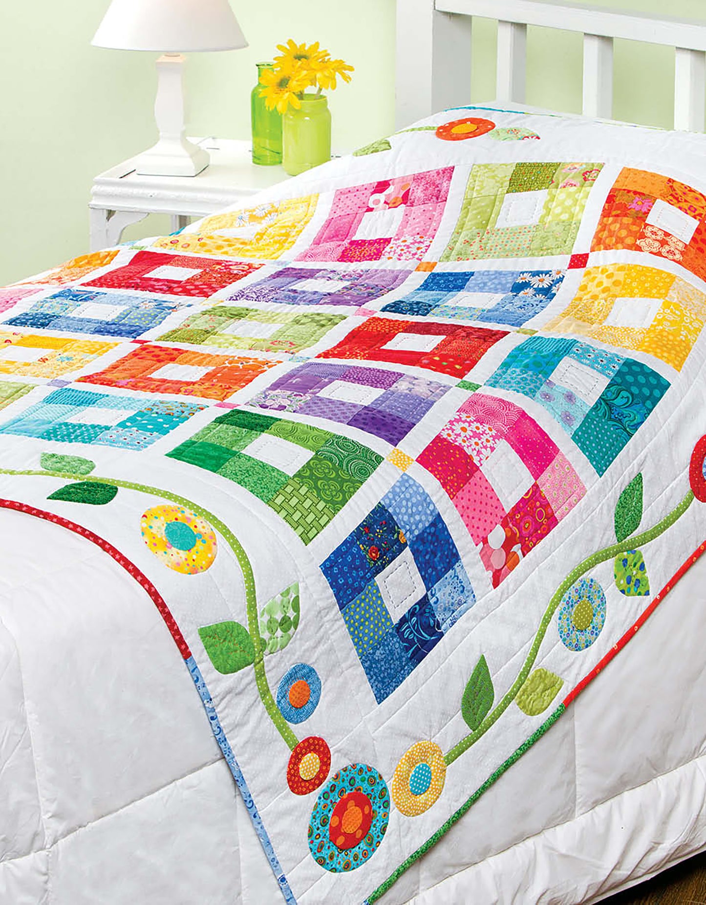 The white bed is covered with Baby Bright Quilts—Use Pops of Color in Stunning Baby Designs! by Leisure Arts, featuring patchwork squares and a floral appliqué border. A lamp and yellow flowers sit on the bedside table.