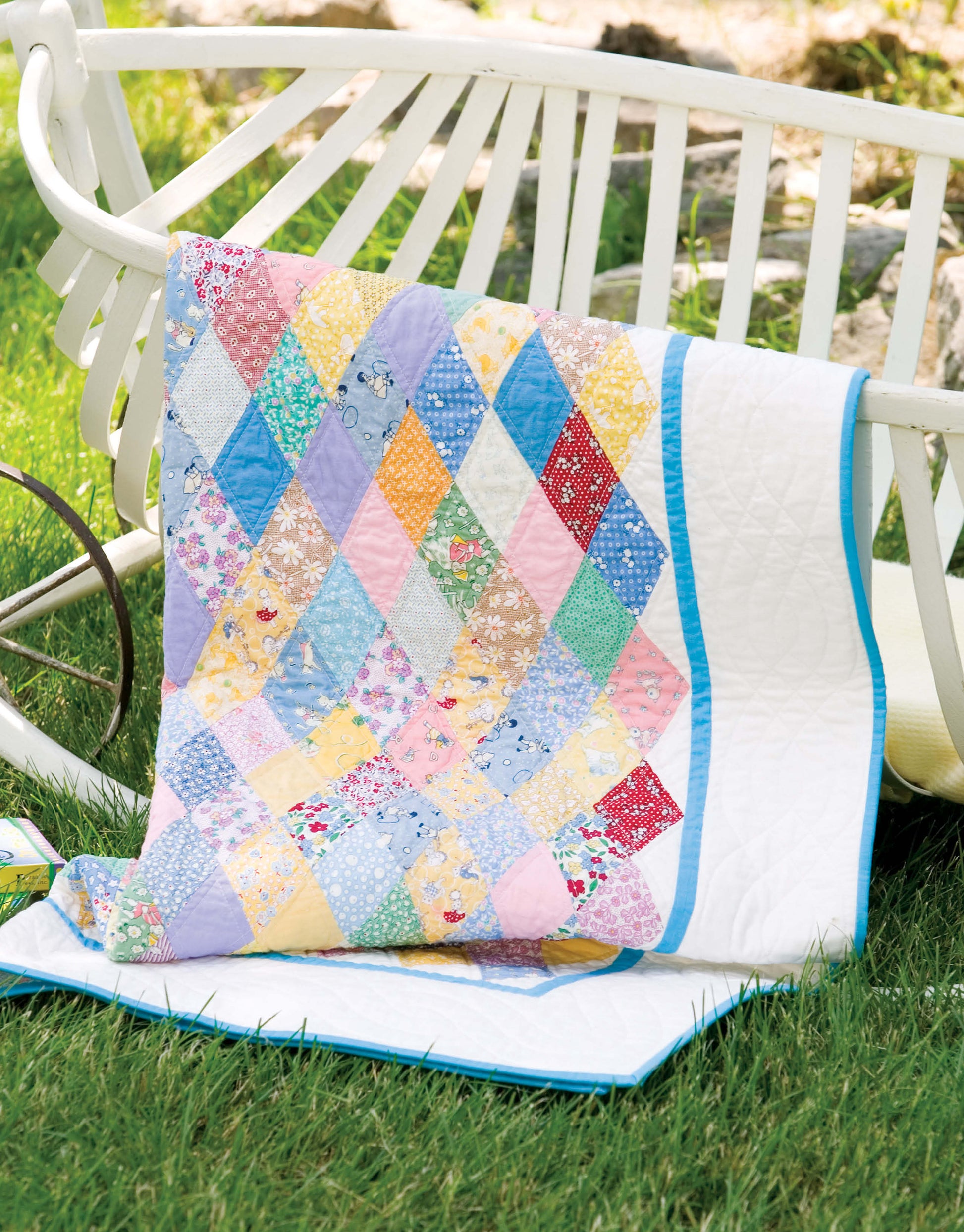 The Baby Bright Quilts by Leisure Arts, featuring vibrant diamond patterns and a blue border, is draped over a white garden bench on a grassy lawn.