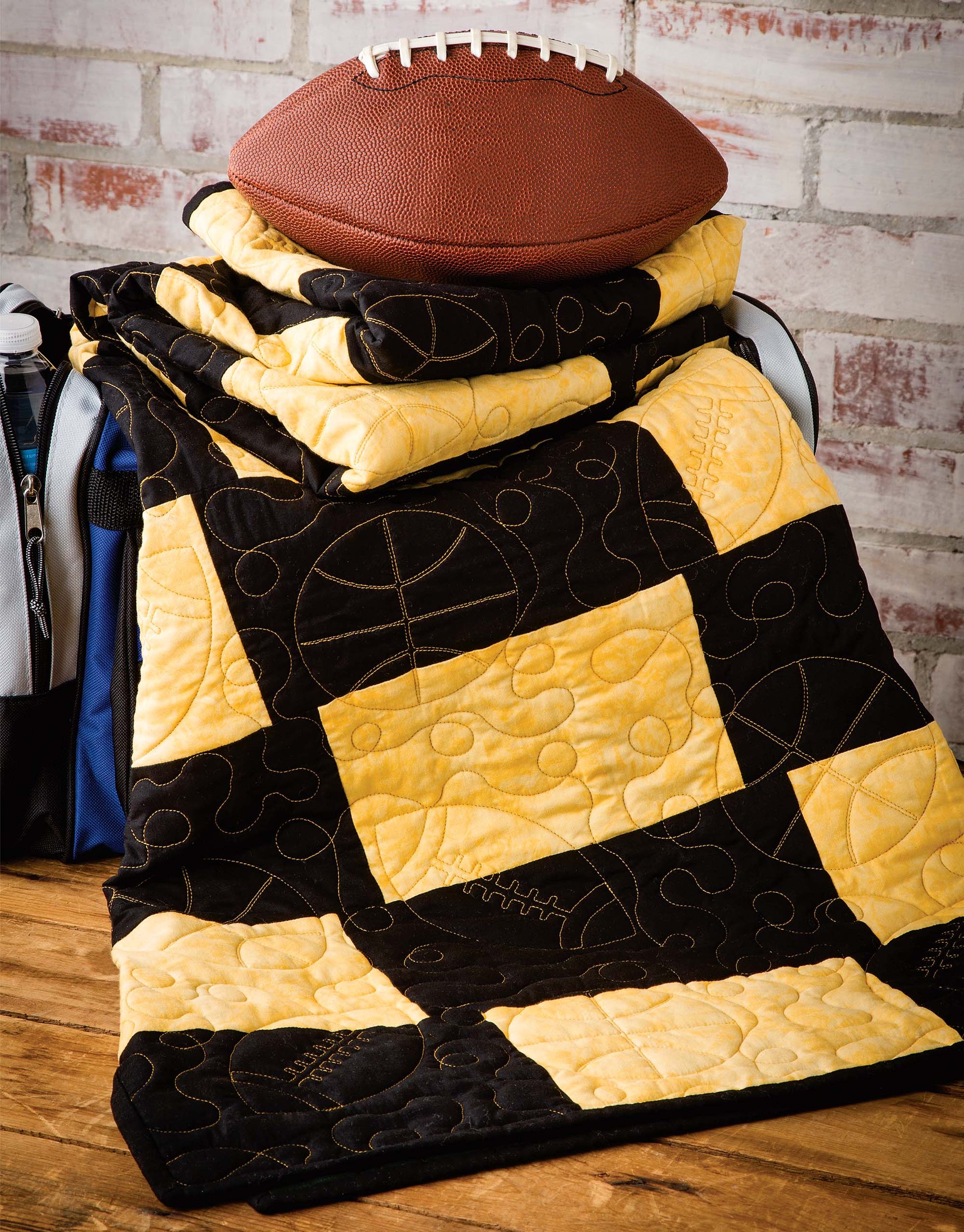 A brown football sits on a black and yellow Quick & Easy Quilts by Leisure Arts, featuring sports-themed stitching, draped over a sports bag with a brick wall background and wooden floor. Perfect inspiration for beginner quilters!.