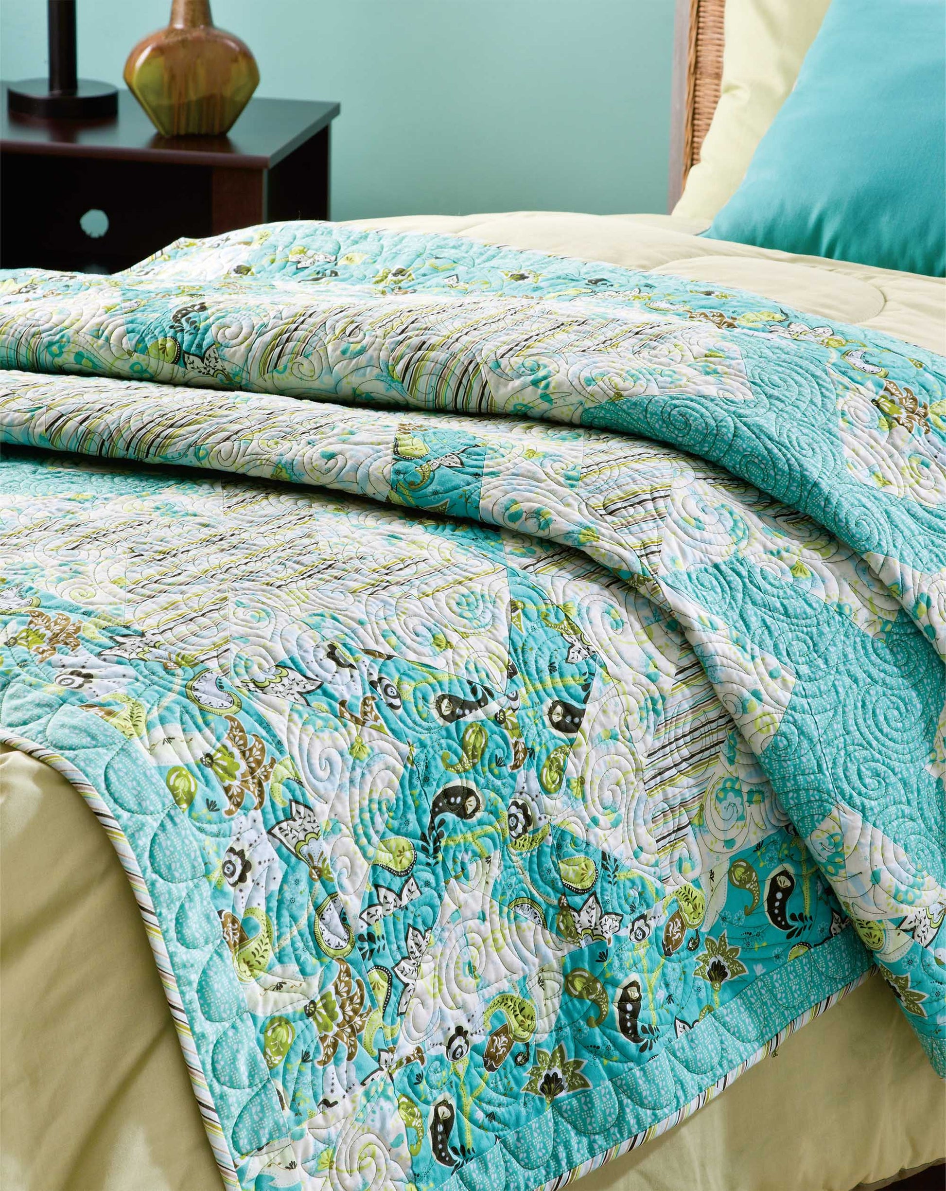 A neatly made bed features a vibrant quilt, created from Leisure Arts' Quick & Easy Quilts—great for fat quarter or beginner projects. At the foot of the bed sits the folded quilt, with a lamp and matching pillow seen on the nightstand behind.