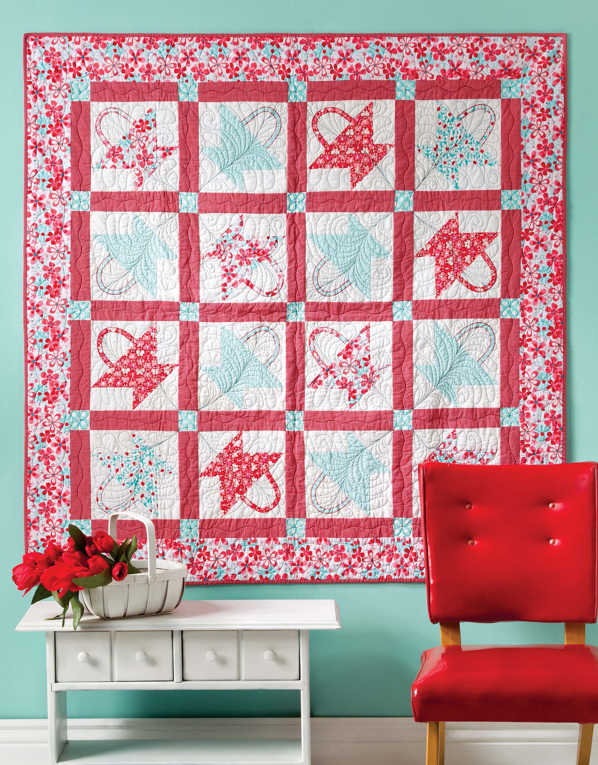 The Lap & Throw Quilts by Leisure Arts, featuring vibrant pink, red, and blue patchwork, is displayed on a teal wall above a white console table with a basket of red flowers and next to a shiny red chair.