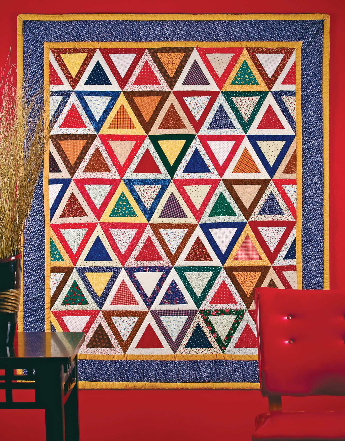 The Leisure Arts Lap & Throw Quilts features rows of multicolored triangles bordered by yellow and blue, displayed on a bright red wall with a red chair and black table with a vase of dried grass in the foreground—perfect for beginner projects.