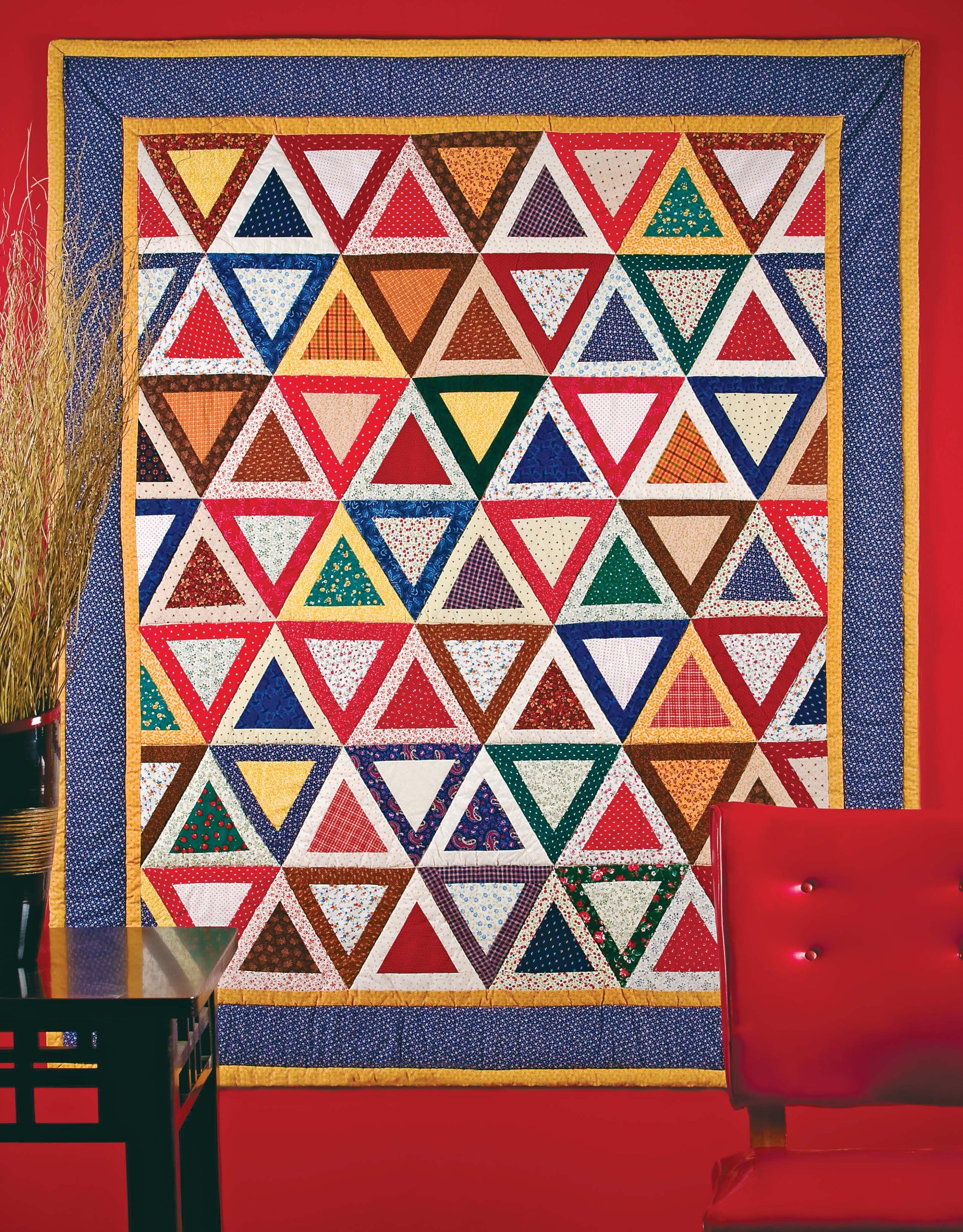 The Leisure Arts Lap & Throw Quilts features rows of multicolored triangles bordered by yellow and blue, displayed on a bright red wall with a red chair and black table with a vase of dried grass in the foreground—perfect for beginner projects.