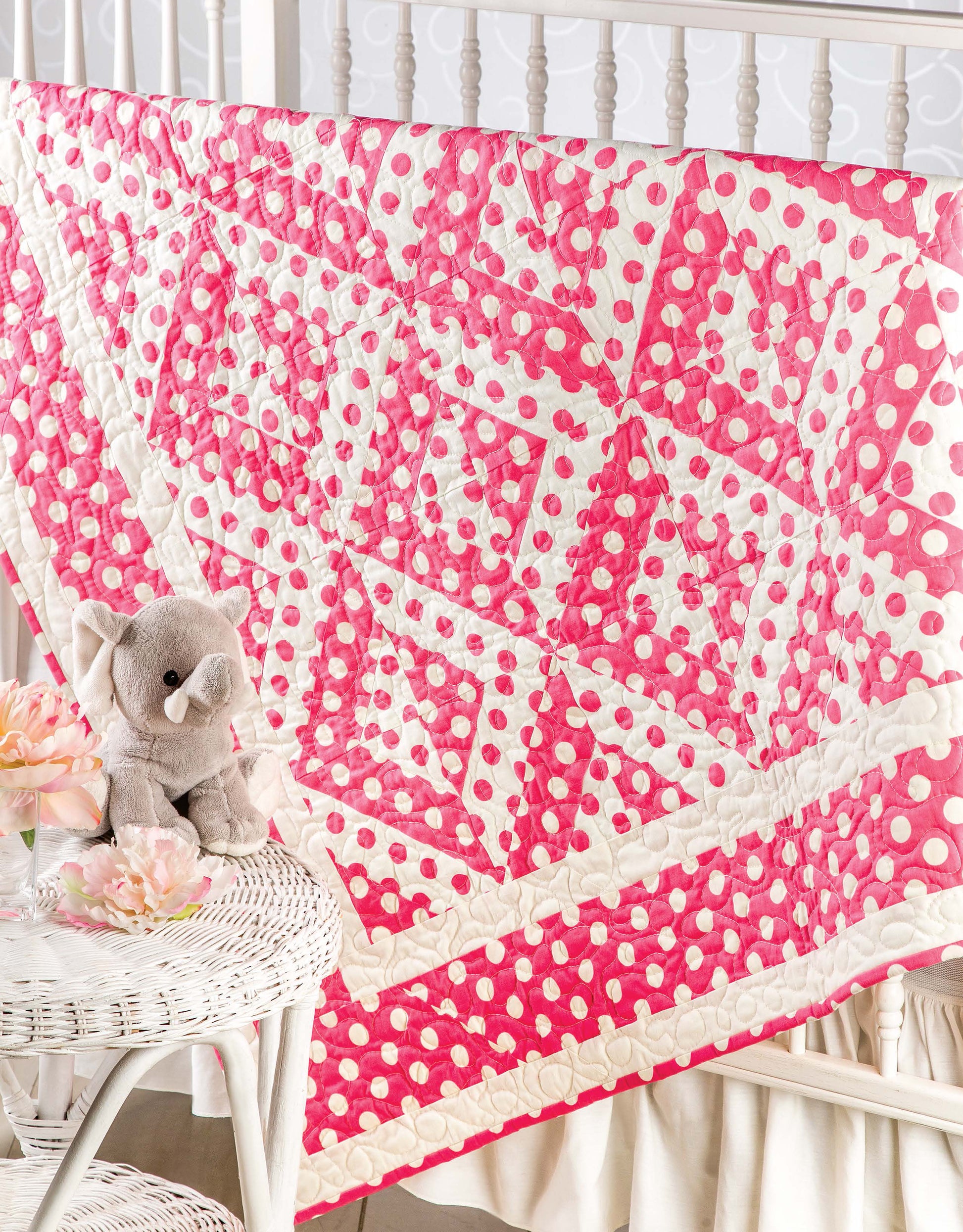 A Lap & Throw Quilt by Leisure Arts in pink and white polka dots drapes over a white crib, with a gray stuffed elephant and pink artificial flowers on a white wicker table in the foreground—ideal for beginners or any occasion.