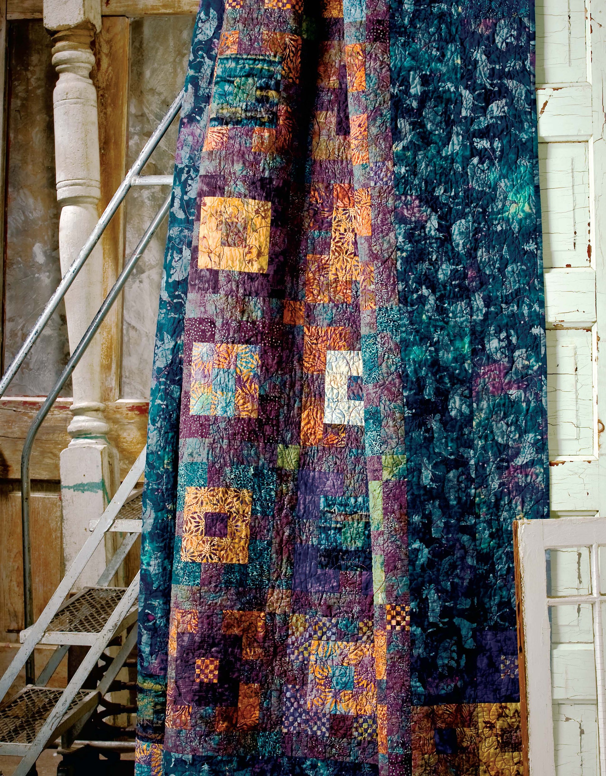 The Lap & Throw Quilts by Leisure Arts, featuring blue, purple, yellow, and orange geometric patterns, hangs beside a metal staircase and rustic wooden railing in a vintage interior.
