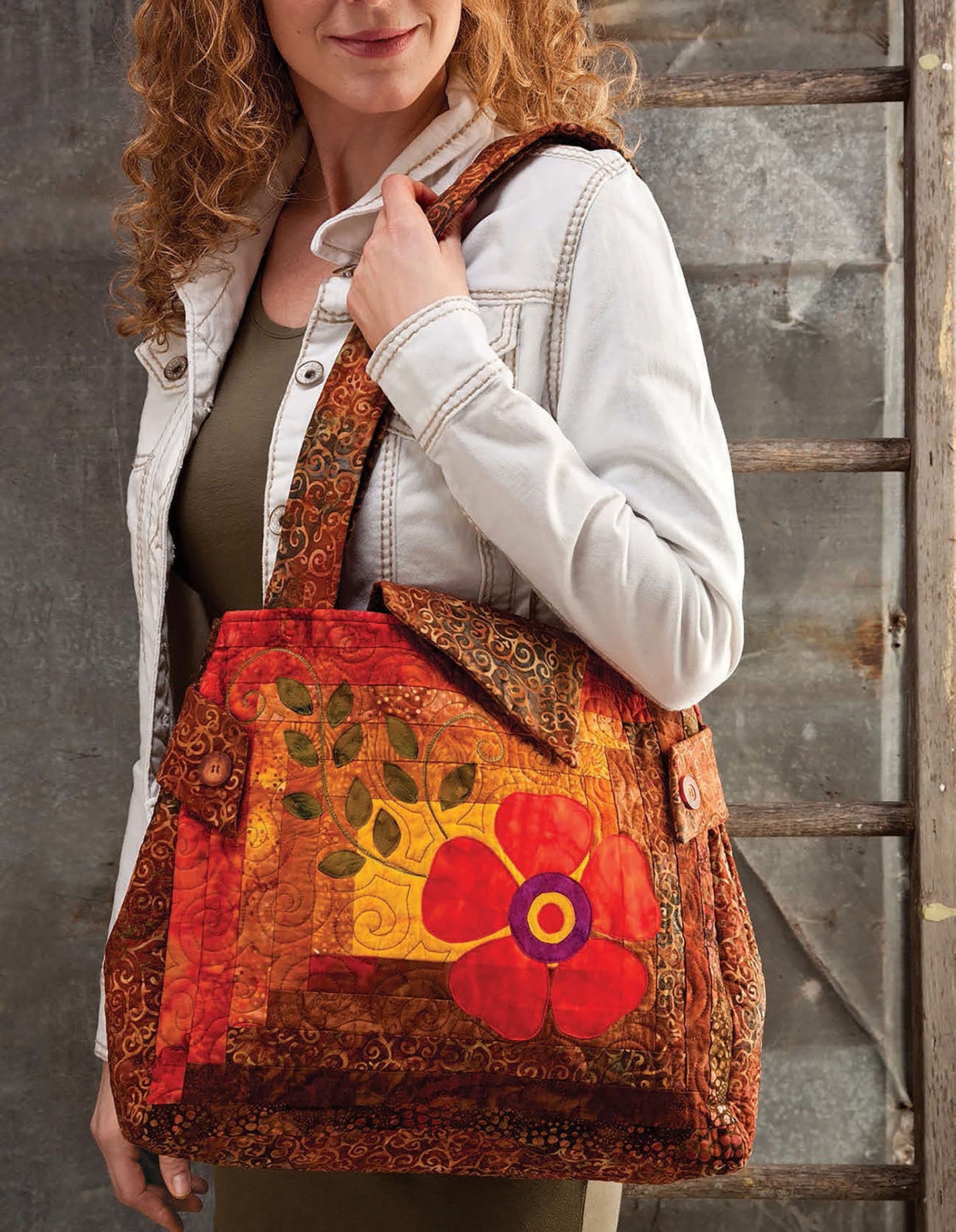 A woman with curly hair in a white jacket holds a colorful Leisure Arts Quilted Purses & Totes handbag with a large red flower applique, standing beside a wooden ladder and rustic wall.