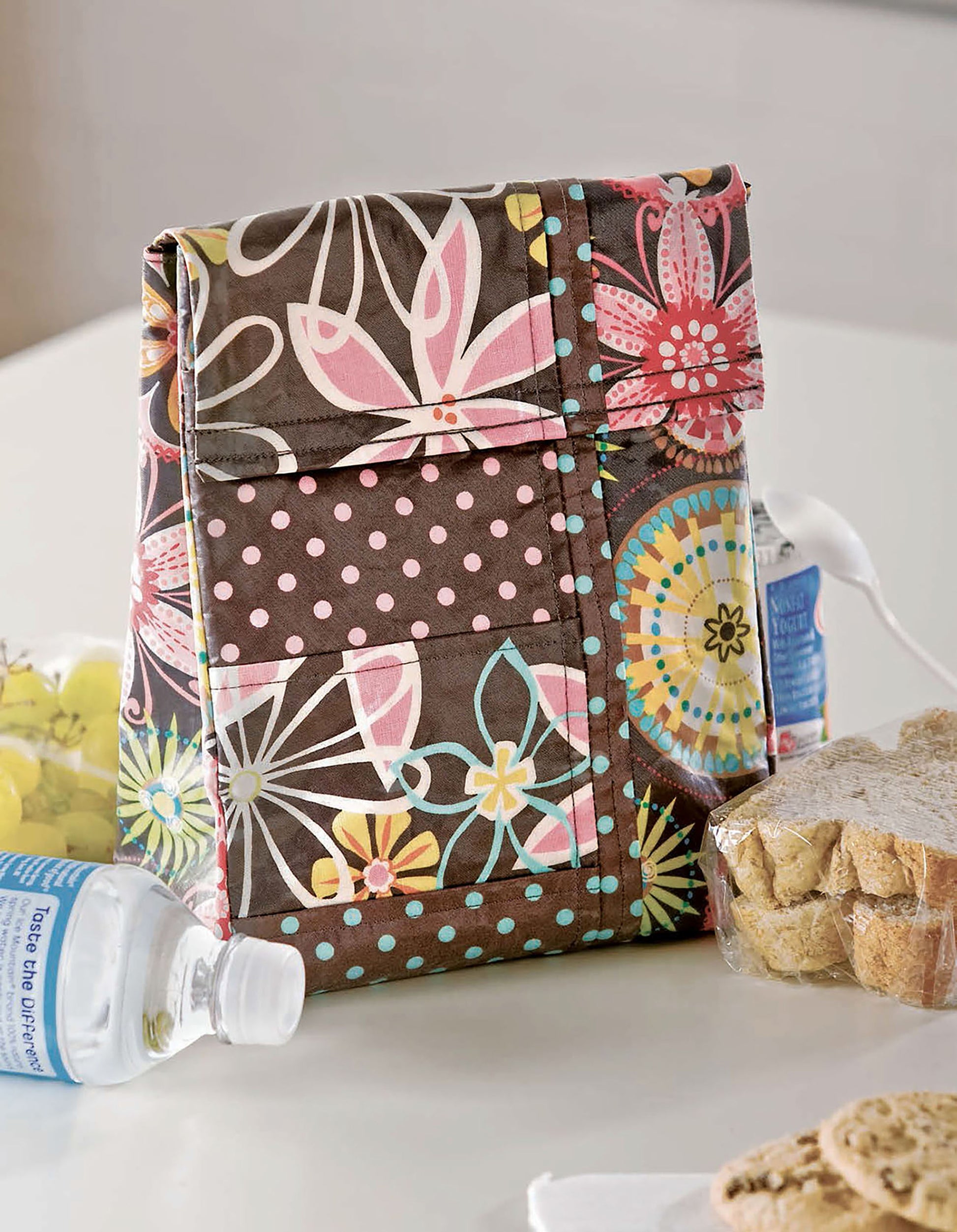 A vibrant, floral-patterned Quilted Purses & Totes lunch bag by Leisure Arts sits on a table beside a water bottle, grapes, and a wrapped sandwich.