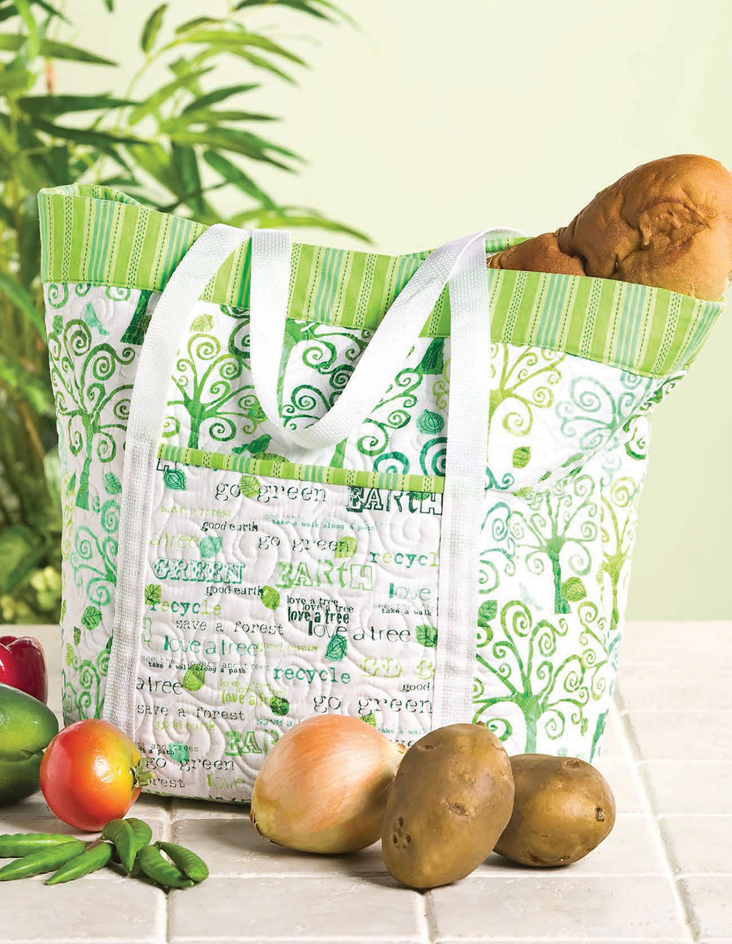 A stylish Quilted Purses & Totes bag by Leisure Arts, featuring green and white patterns, rests on a counter with bread inside and apples, potatoes, and limes around it. A leafy plant in the background highlights its chic everyday design.