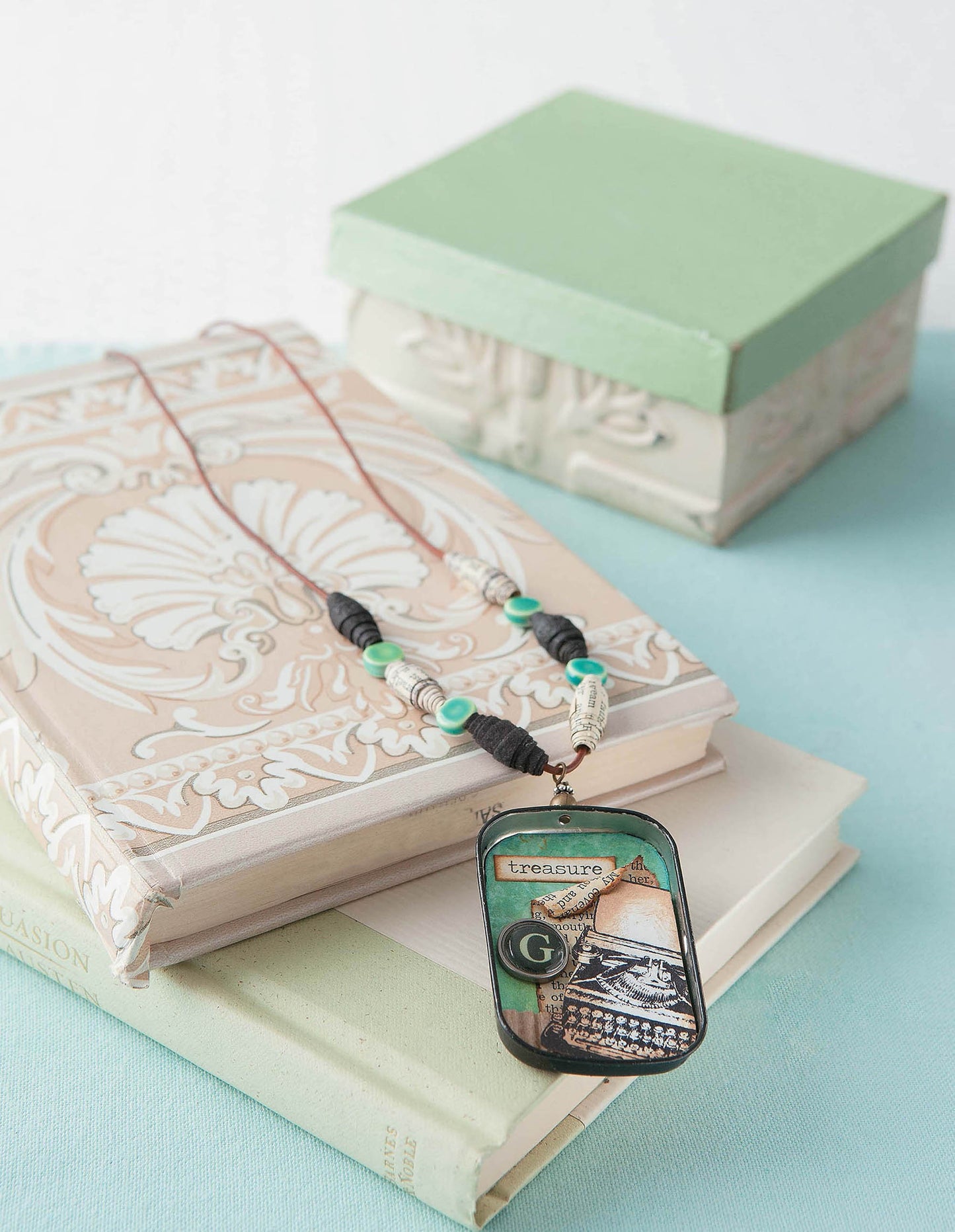 Easy Vintage Jewelry from Leisure Arts features a decorative necklace with green and black beads and a vintage typewriter pendant reading "treasure," displayed on ornate books beside a green box on a pale blue surface.