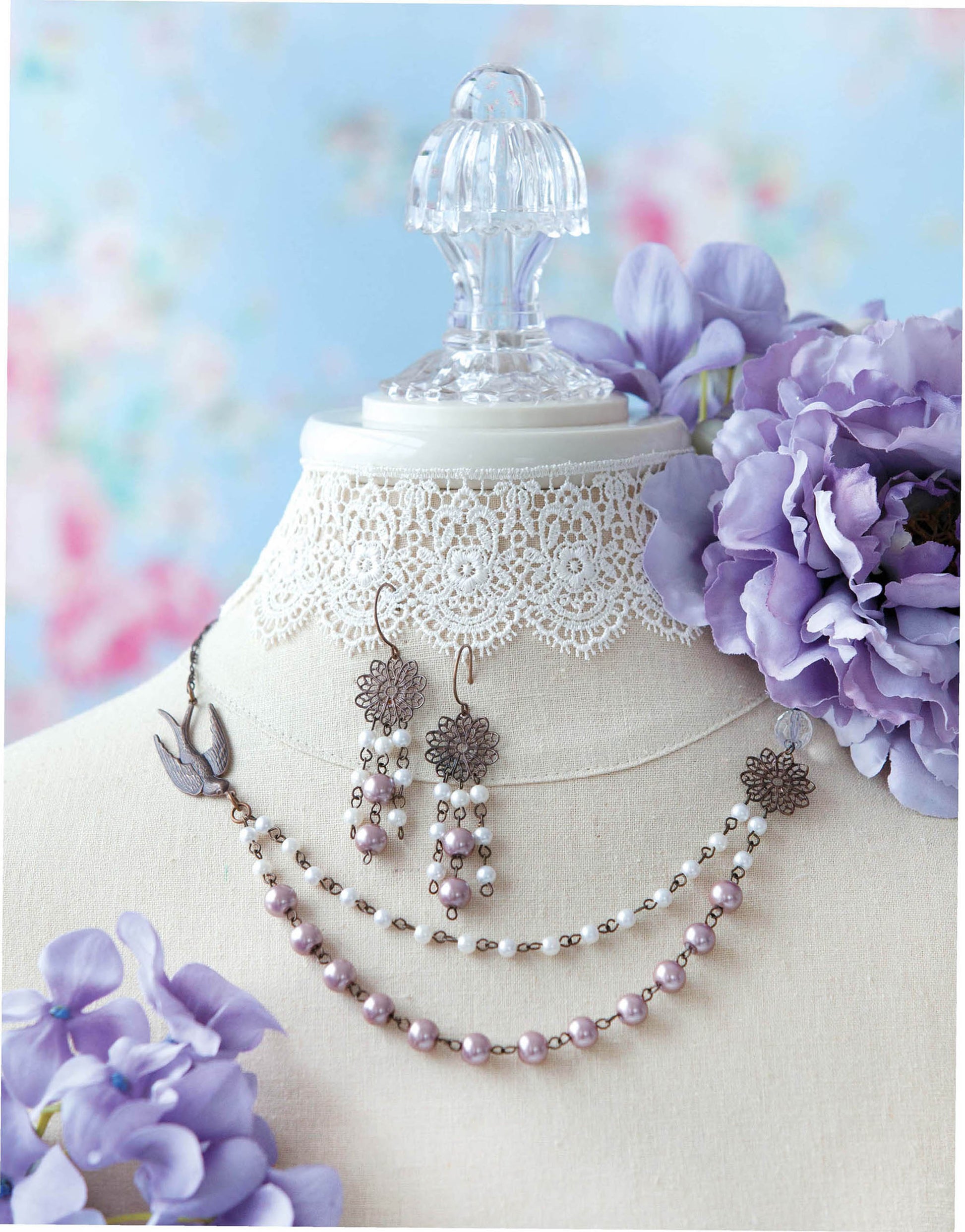 The Easy Vintage Jewelry set by Leisure Arts features a lace-adorned display bust showcasing a purple beaded necklace and matching earrings with floral accents, arranged among purple flowers on a soft blue background.