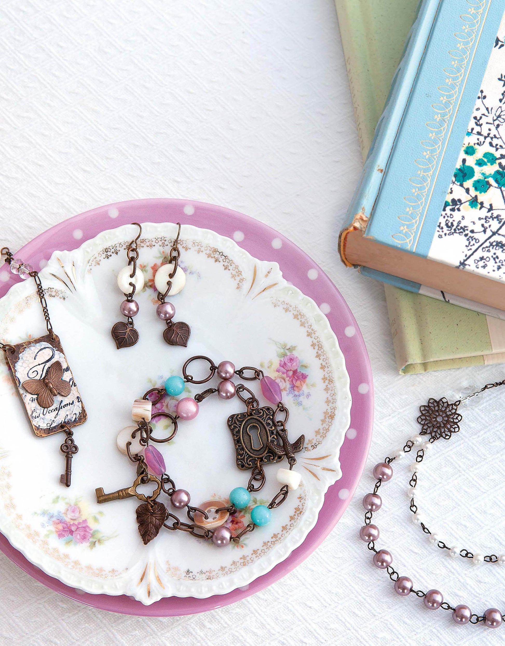 Easy Vintage Jewelry by Leisure Arts features pearls, charms, and keys on a pink plate. Perfect for vintage jewelry crafts enthusiasts, it’s paired with a beaded bracelet and ornate blue and green books.