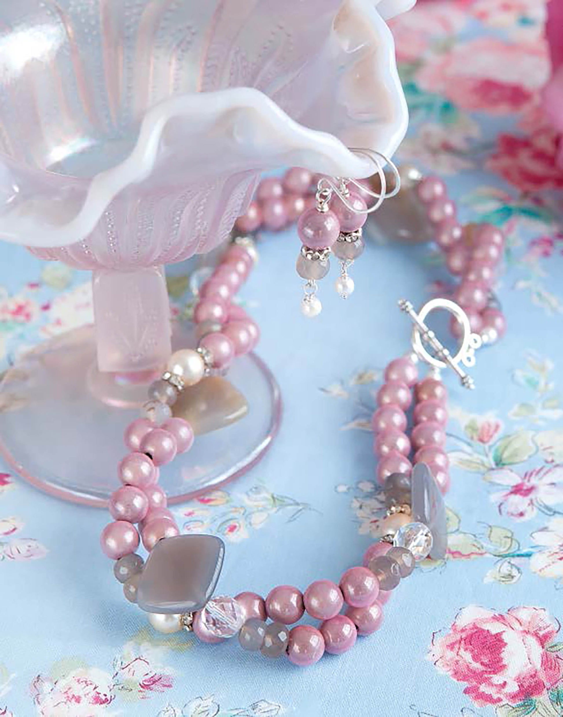 Easy Vintage Jewelry by Leisure Arts—a pink pearl beaded necklace and matching earrings for beginner jewelry makers—rests beside a pink glass dish on light blue floral fabric with silver accents.
